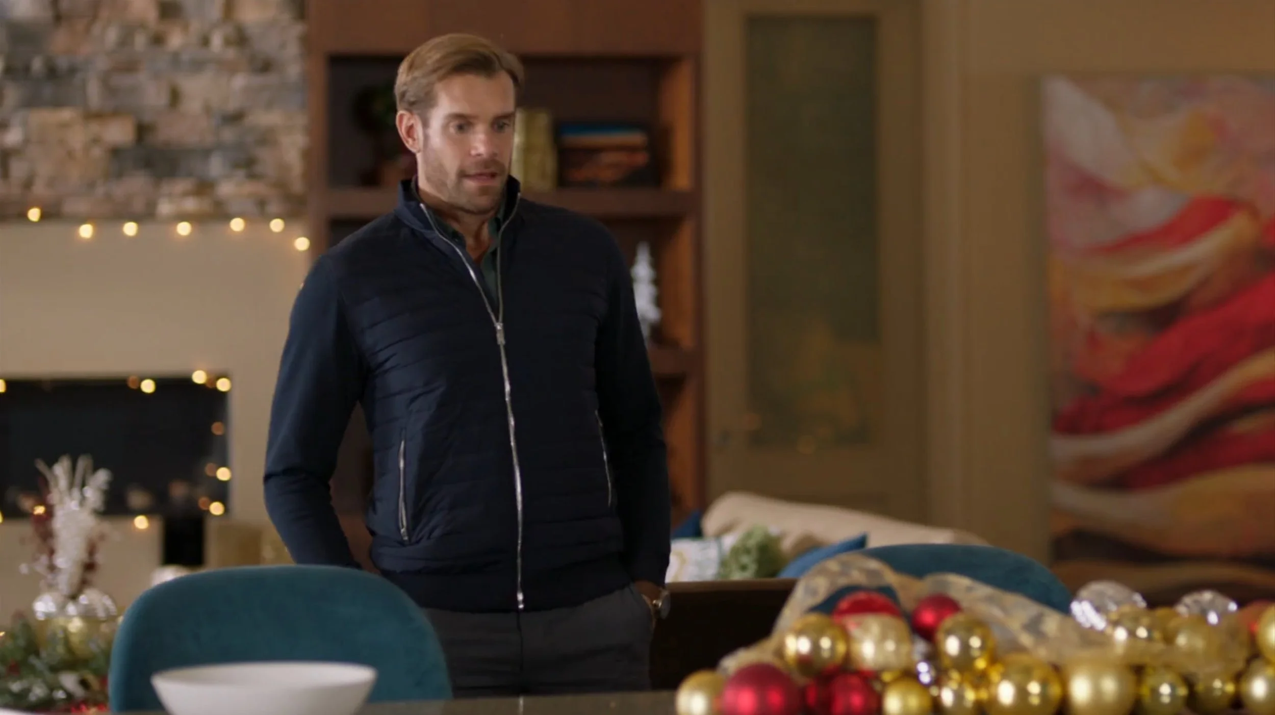 A man with light brown hair and a beard, wearing a dark zippered jacket, stands in a decorated living room with Christmas ornaments on the table in front of him.