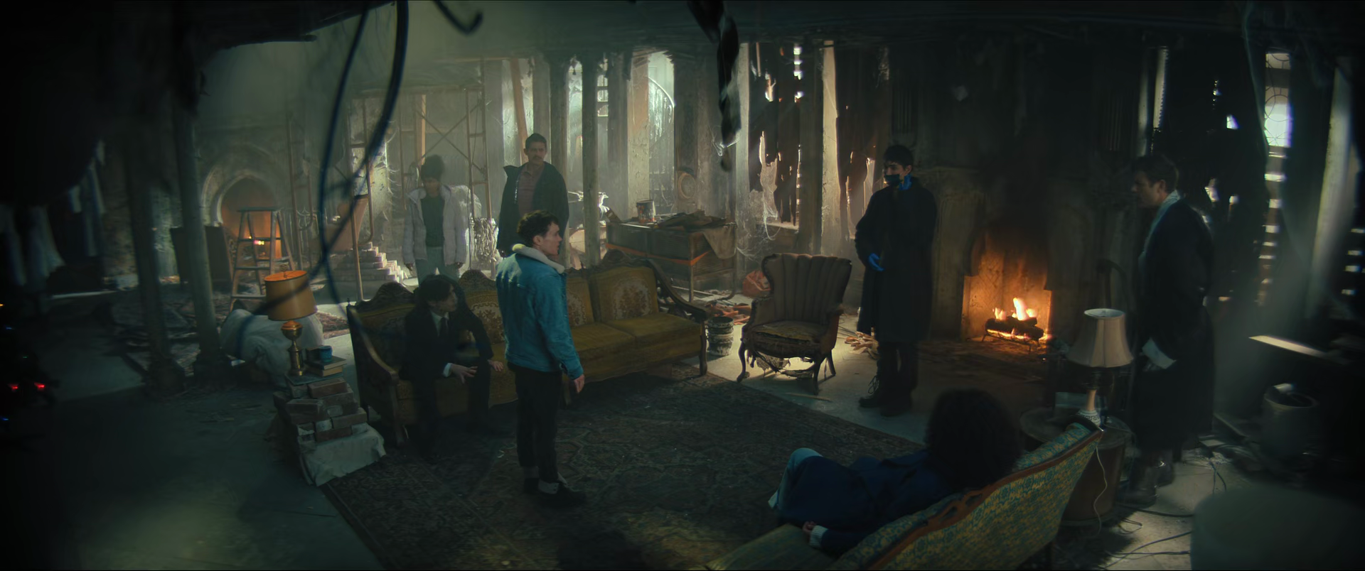 Six people gathered in a dimly lit, cluttered room with a fireplace, old furniture, and scattered objects, creating a mysterious atmosphere.