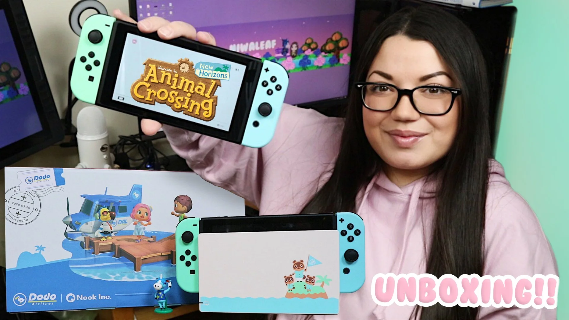 A woman with long dark hair and glasses is smiling while holding a Nintendo Switch with Animal Crossing on the screen in front of her. There is a boxed Animal Crossing set on the table, and another Nintendo Switch in front of it with an Animal Crossi