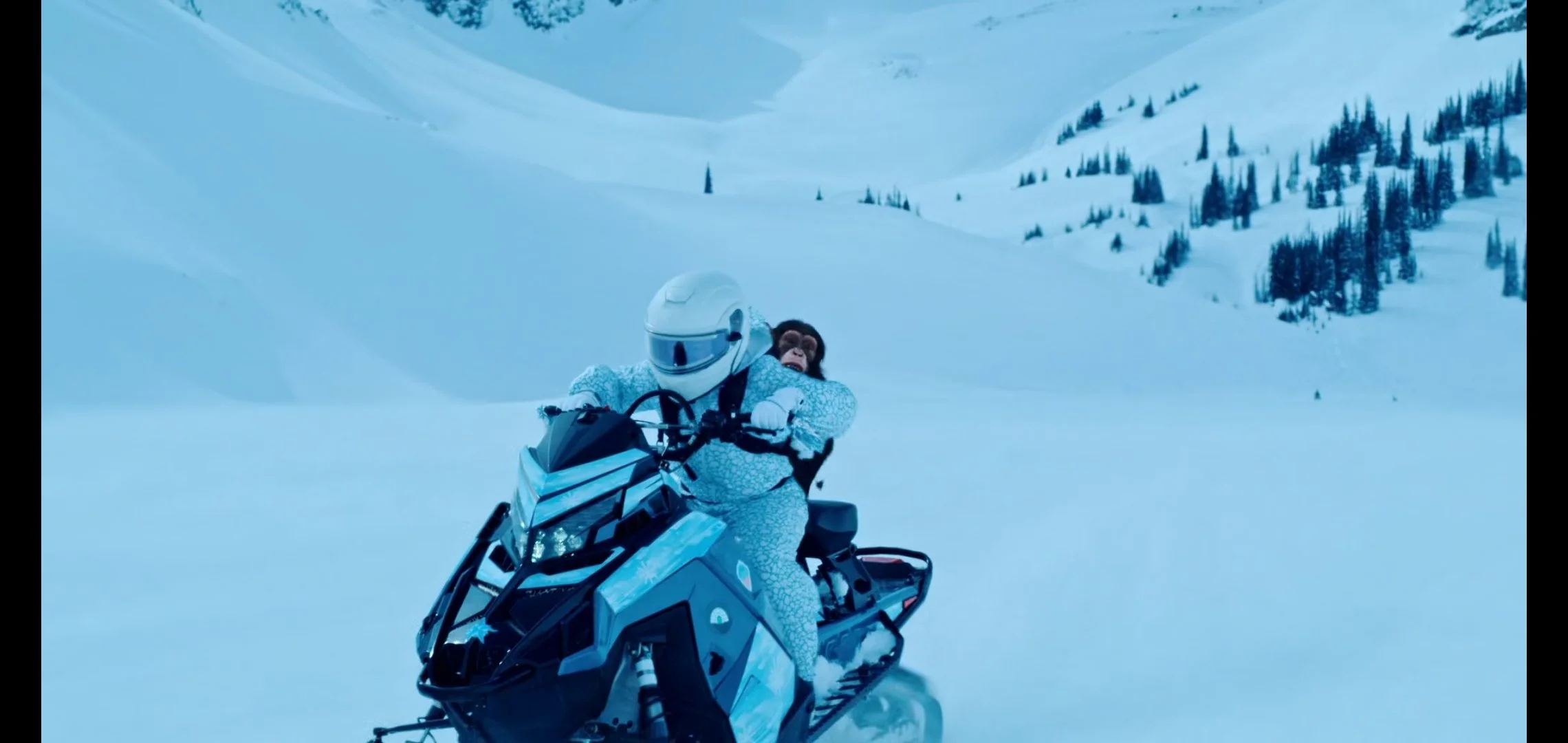 Two people riding a snowmobile on snowy terrain with a mountainous landscape and pine trees in the background.