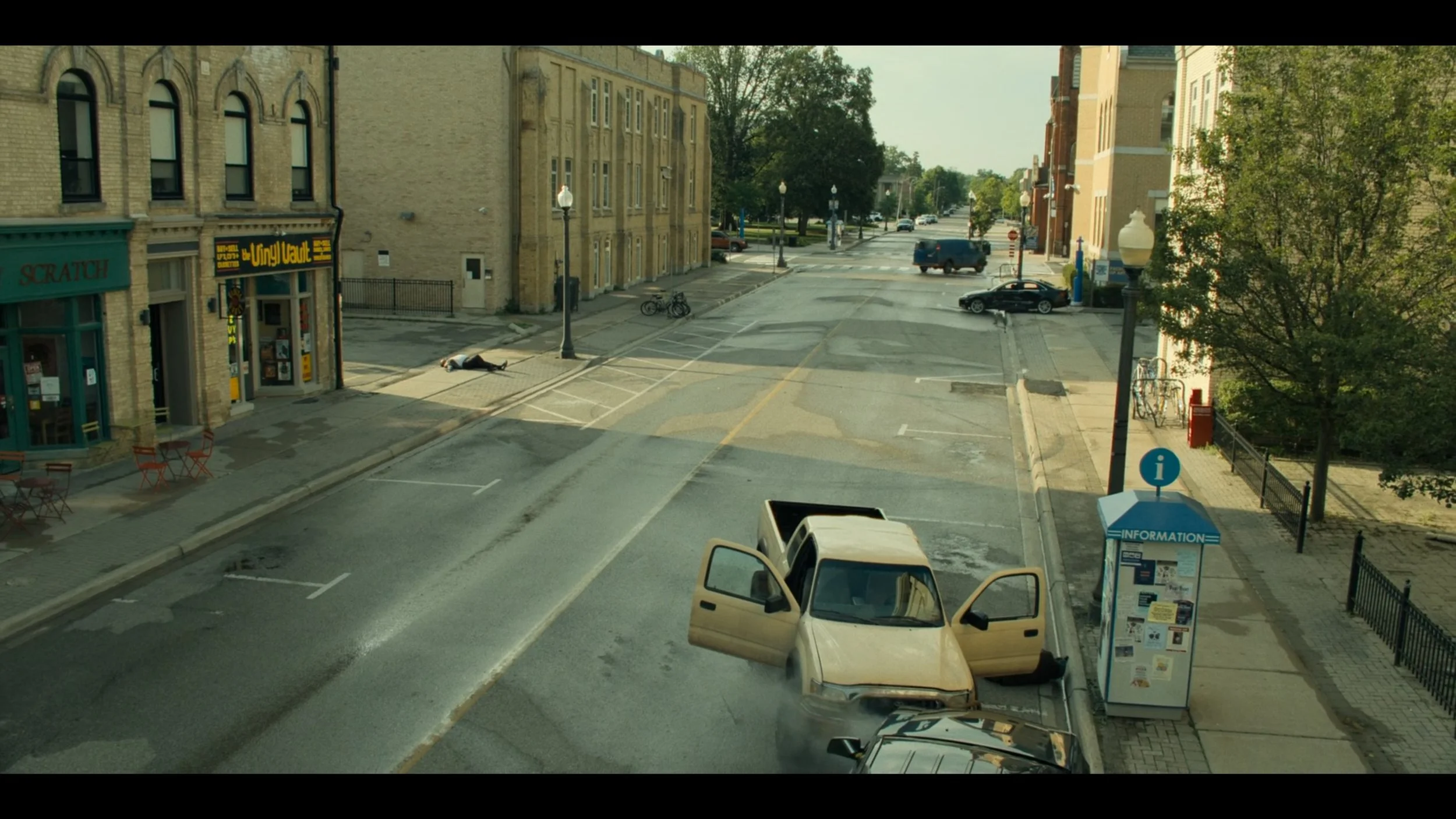 A beige SUV has crashed into a parking meter at an intersection, with its driver’s side door open. There is smoke coming from the front of the SUV. Nearby, a person lies motionless on the sidewalk, and other cars are visible on the street.