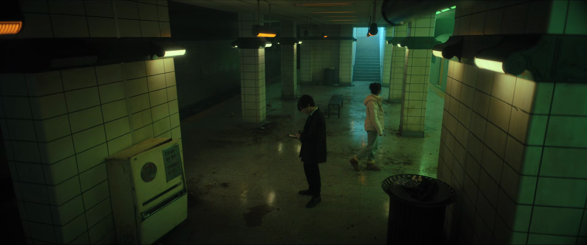 Underground subway station with tiled walls, two people standing, one on their phone and one walking, dim lighting, greenish hue, stairway in the background.