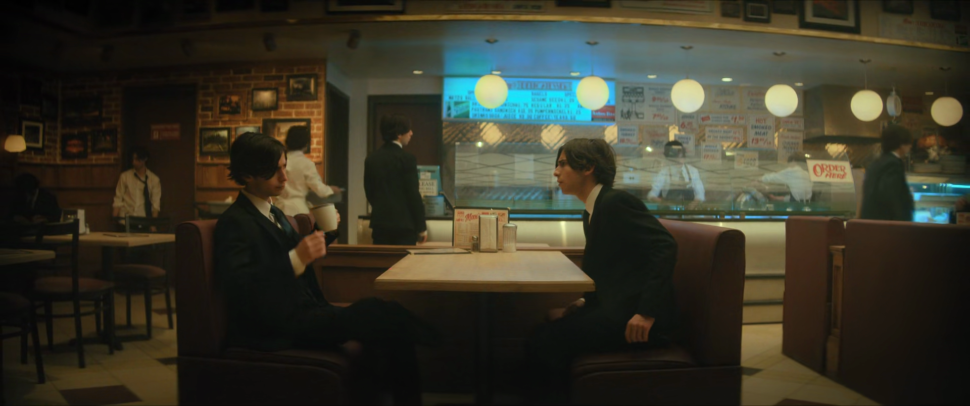 Two people in suits sitting at a booth in a retro-style diner, having a conversation, with a diner counter and staff in the background.
