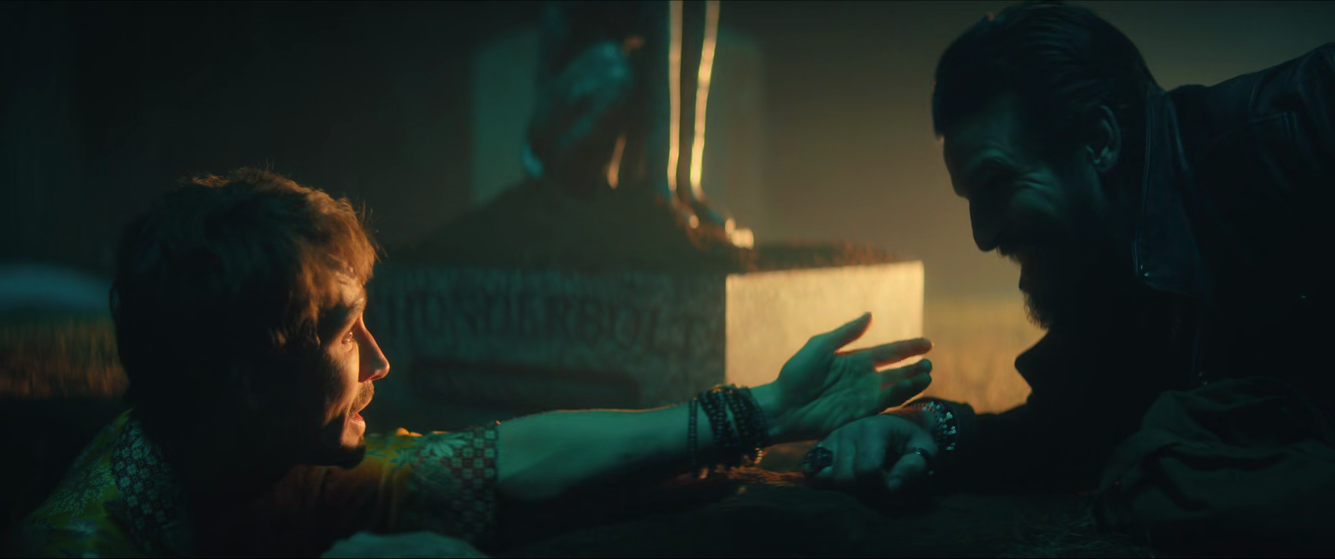 Two men are on the ground, one reaching out to the other in a dark, intense scene with dramatic lighting, with a statue or sculpture visible in the background.