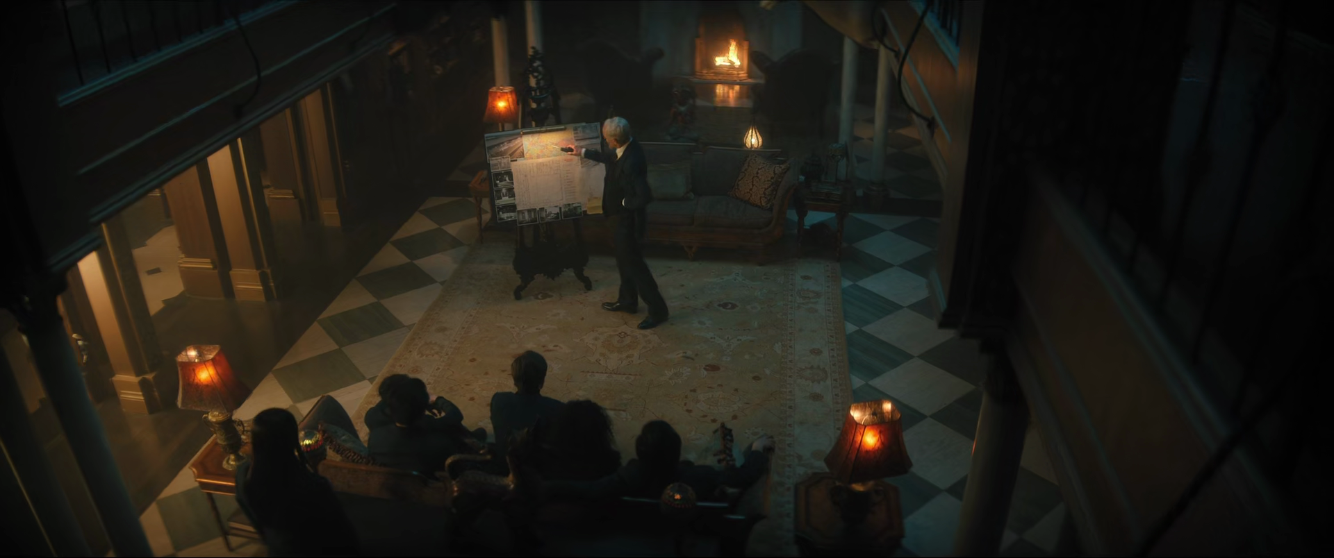 An older man presents a large map on a board in a luxurious living room while four people sit on a sofa watching him.