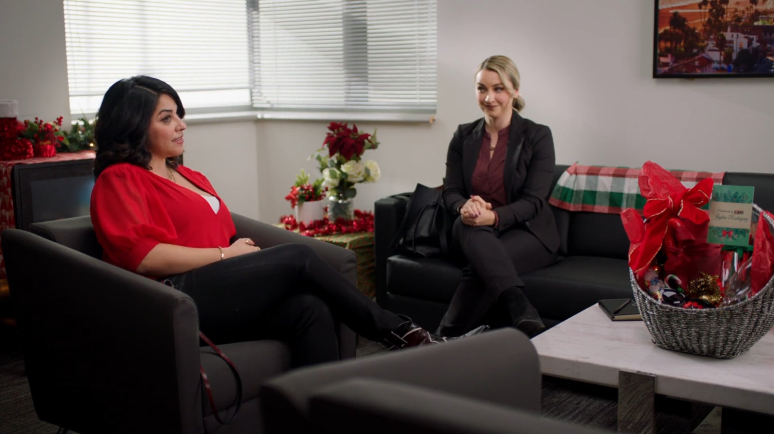 Two women sitting on black sofas in an office decorated for Christmas, with poinsettias, a gift basket, and holiday decorations on the table and in the background.