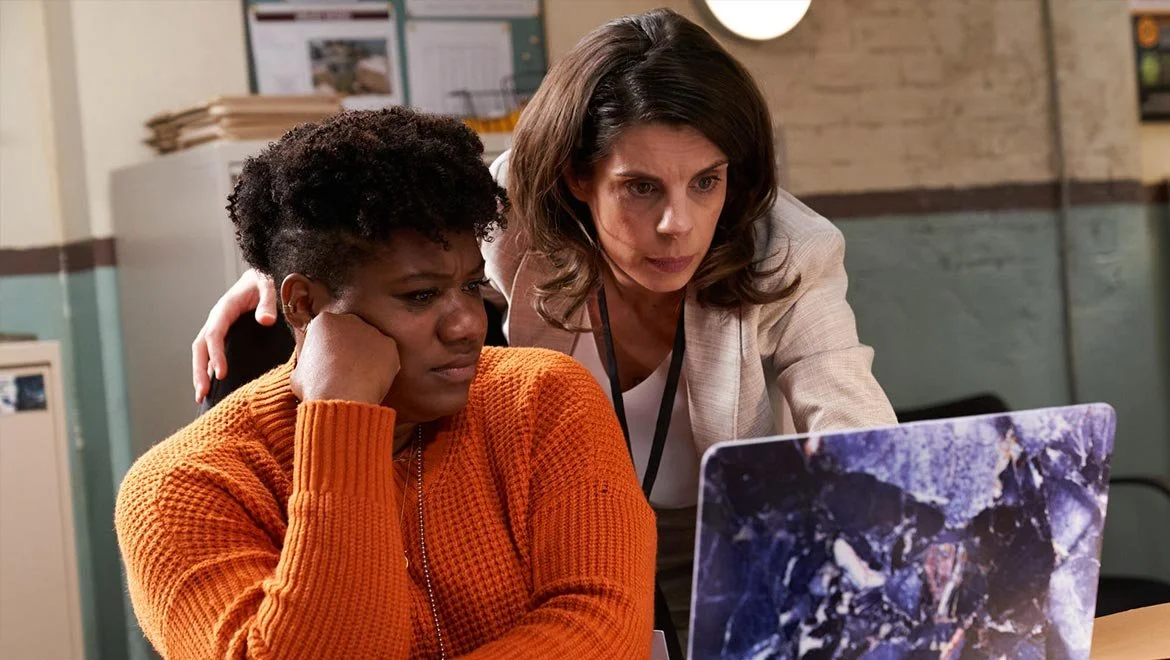 Two women look at a laptop screen with serious expressions; one is sitting and resting her head on her hand, the other is leaning in closely.