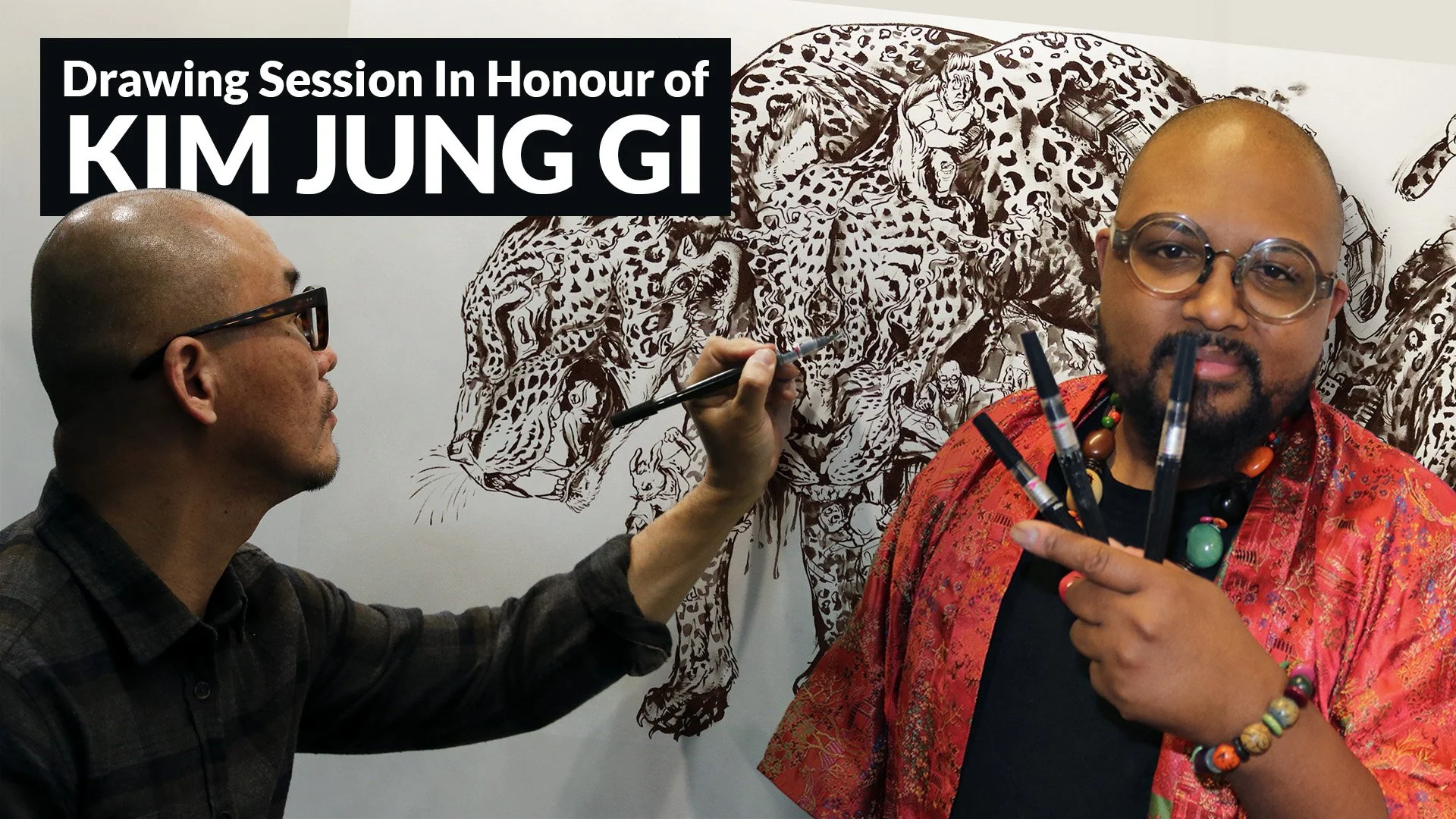 Two artists creating a detailed drawing of a roaring lion on a white wall. One artist is bald, wearing glasses and a plaid shirt, holding a pen. The other artist has a shaved head, glasses, a black shirt, and a colorful necklace, holding several draw