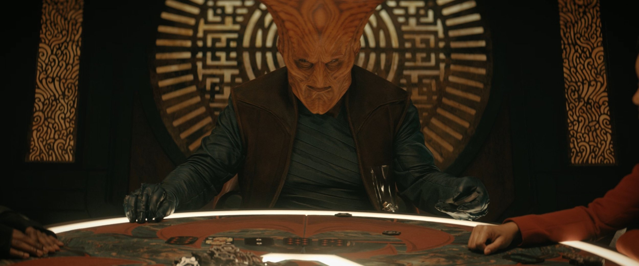A humanoid character with an orange, textured alien face and large forehead, seated at a gaming table with poker chips, against a decorative background with Asian-inspired patterns.