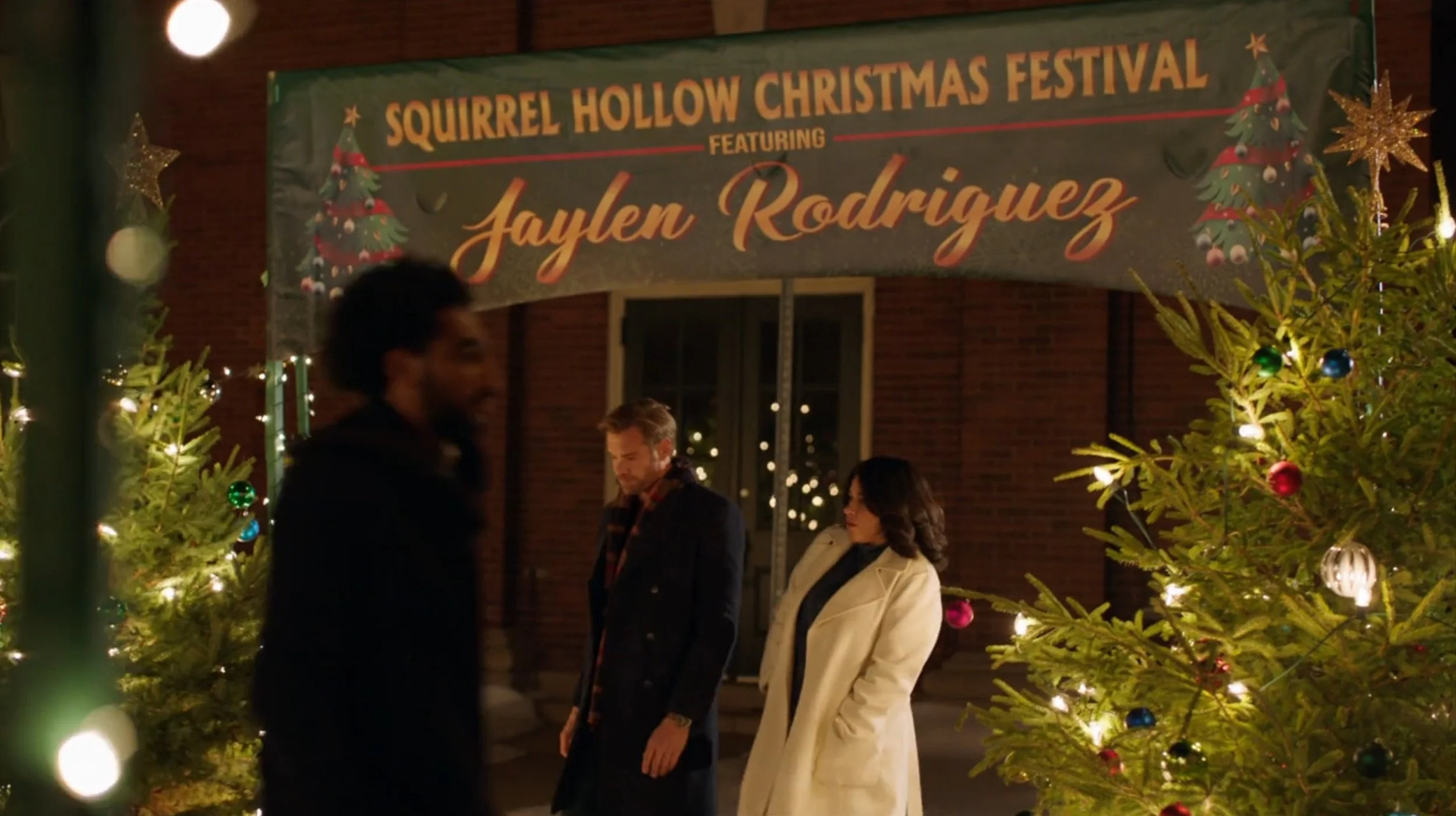 A Christmas festival event titled 'Squirrel Hollow Christmas Festival' featuring Jaylen Rodriguez, with people standing near decorated Christmas trees illuminated with lights and ornaments, outside a brick building at night.