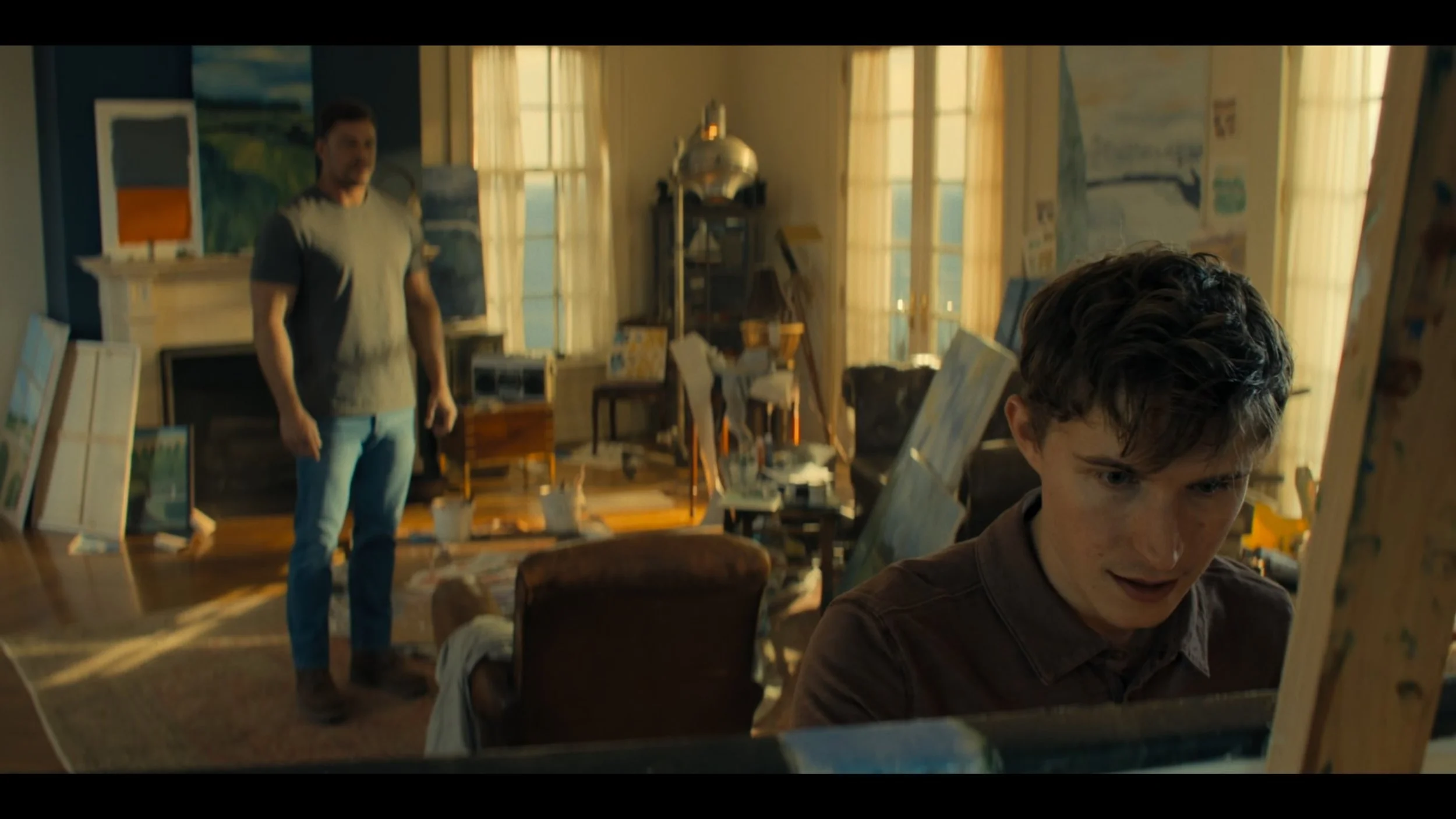 A young man with dark curly hair painting on a canvas in an art studio, with an older man standing watching in the background, surrounded by paintings and art supplies.