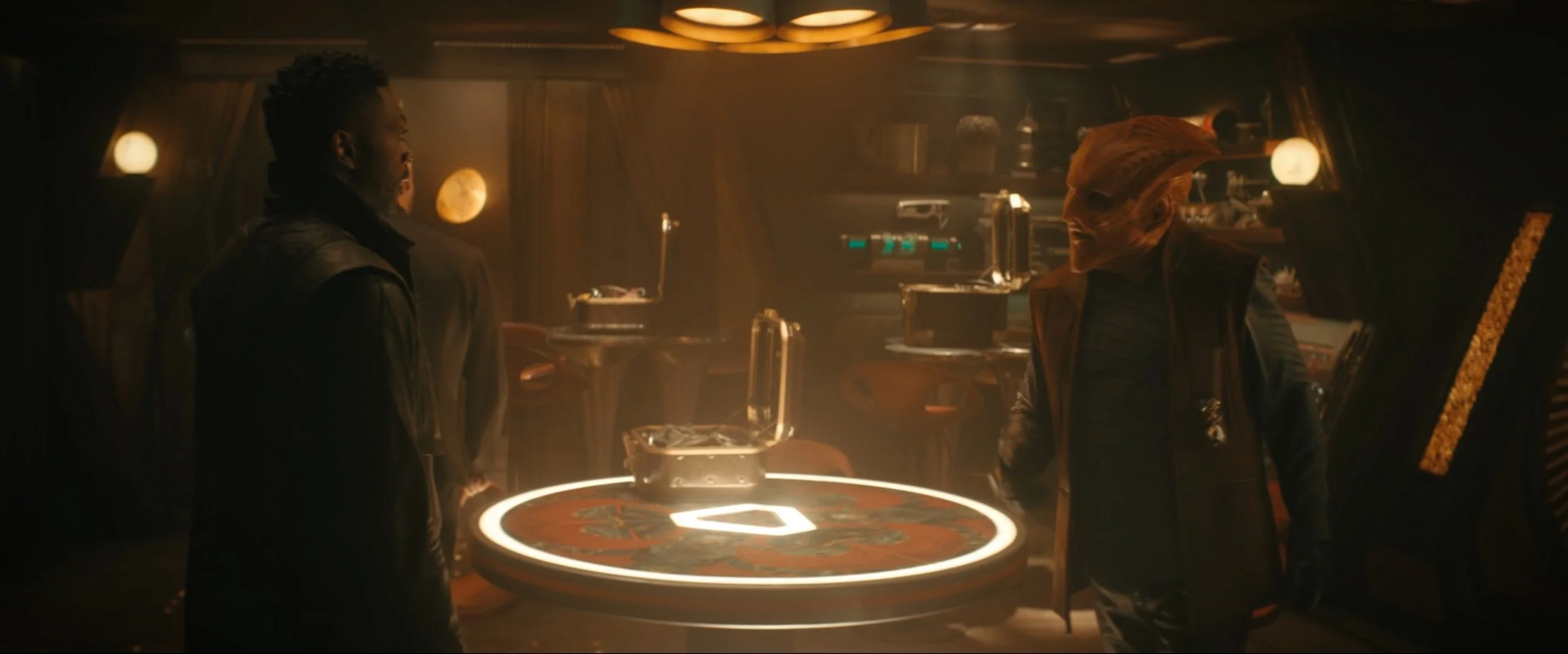 Two men are in a dimly lit room with a circular table in the center, with neon lighting around its edge, and some equipment and shelves in the background.