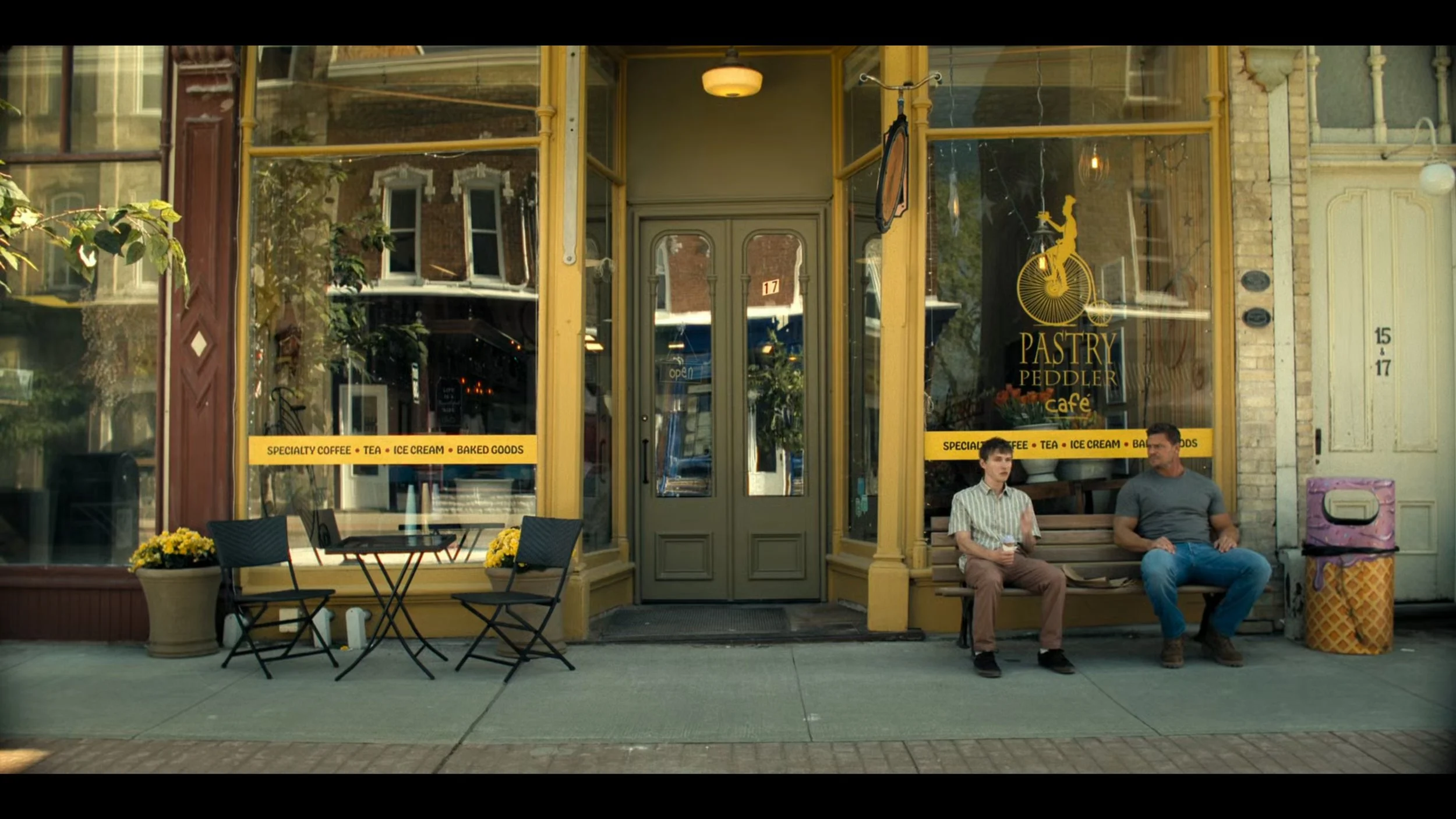 Two men sitting on a bench outside a bakery cafe called Pastry Peddler, with a small outdoor table and chairs, and yellow flowers in pots.