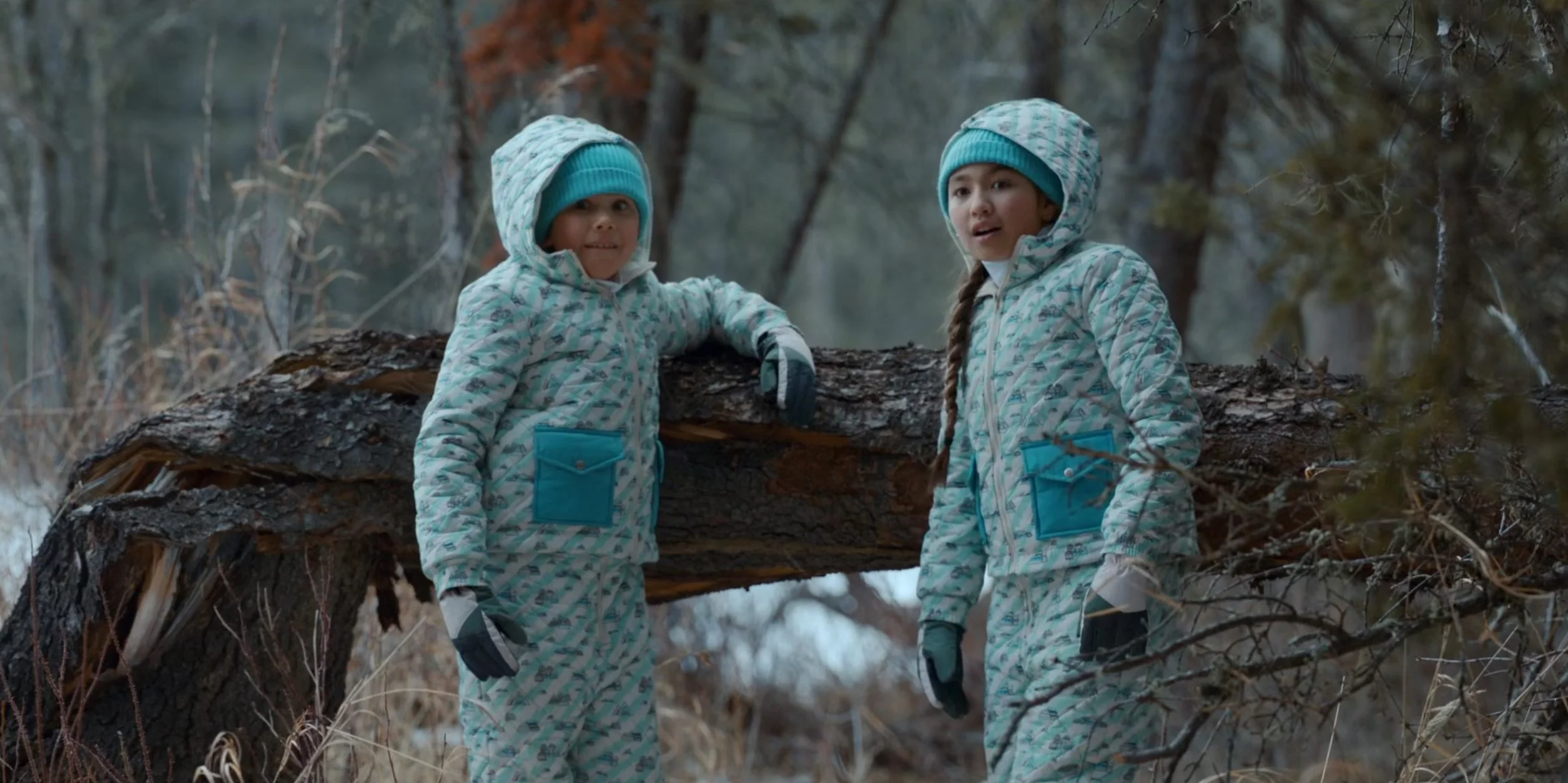Two young girls in winter clothing standing outdoors near a fallen tree in a forest during winter.