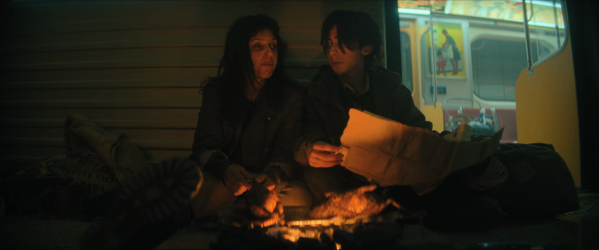 Two people sitting on a sidewalk at night, reading a paper by a small fire, with a yellow food truck with colorful posters in the background.