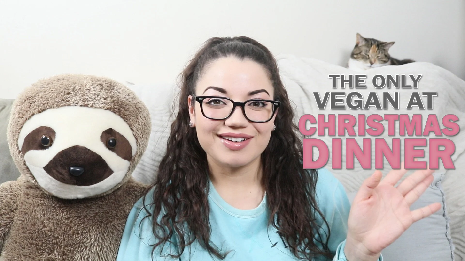 A woman with glasses is sitting on a couch in front of a sloth stuffed animal and a cat resting on a pillow in the background. There is text overlay that says 'The only vegan at Christmas dinner'.