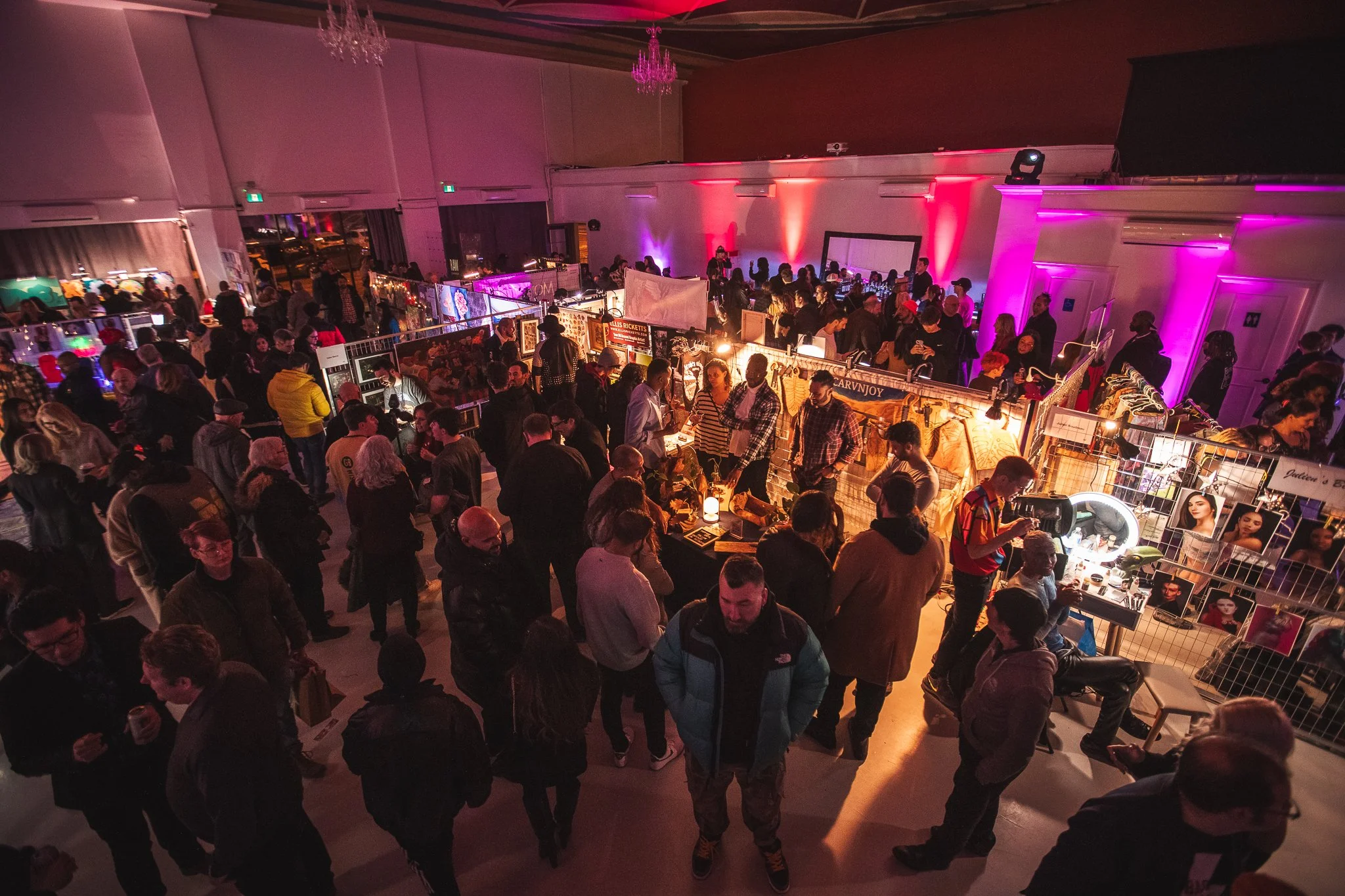 Indoor event with people browsing vendor booths illuminated by colorful lights, with a high ceiling and chandeliers.