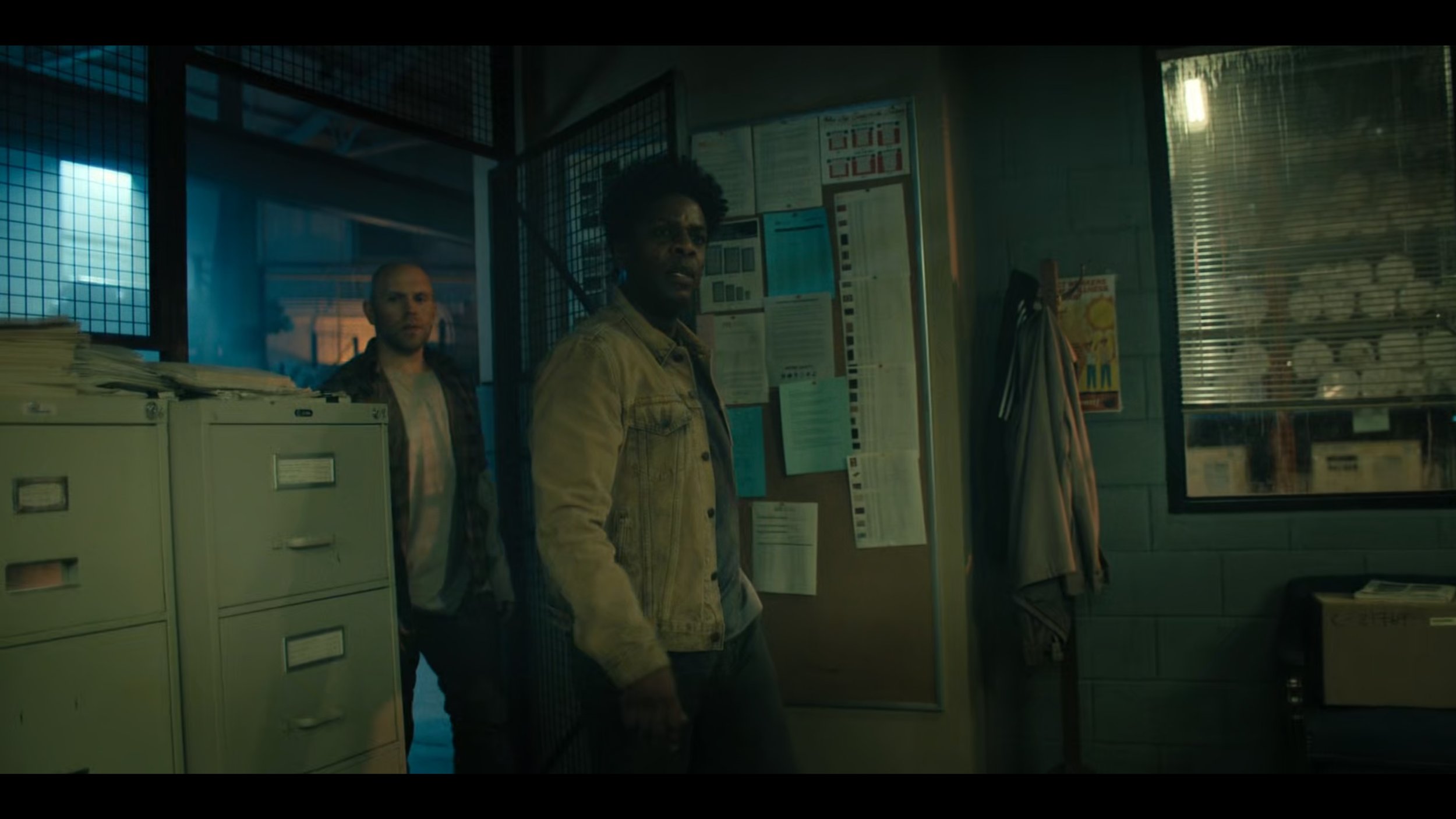 Two men standing in a dimly lit office with file cabinets, bulletin board, and window with blinds.