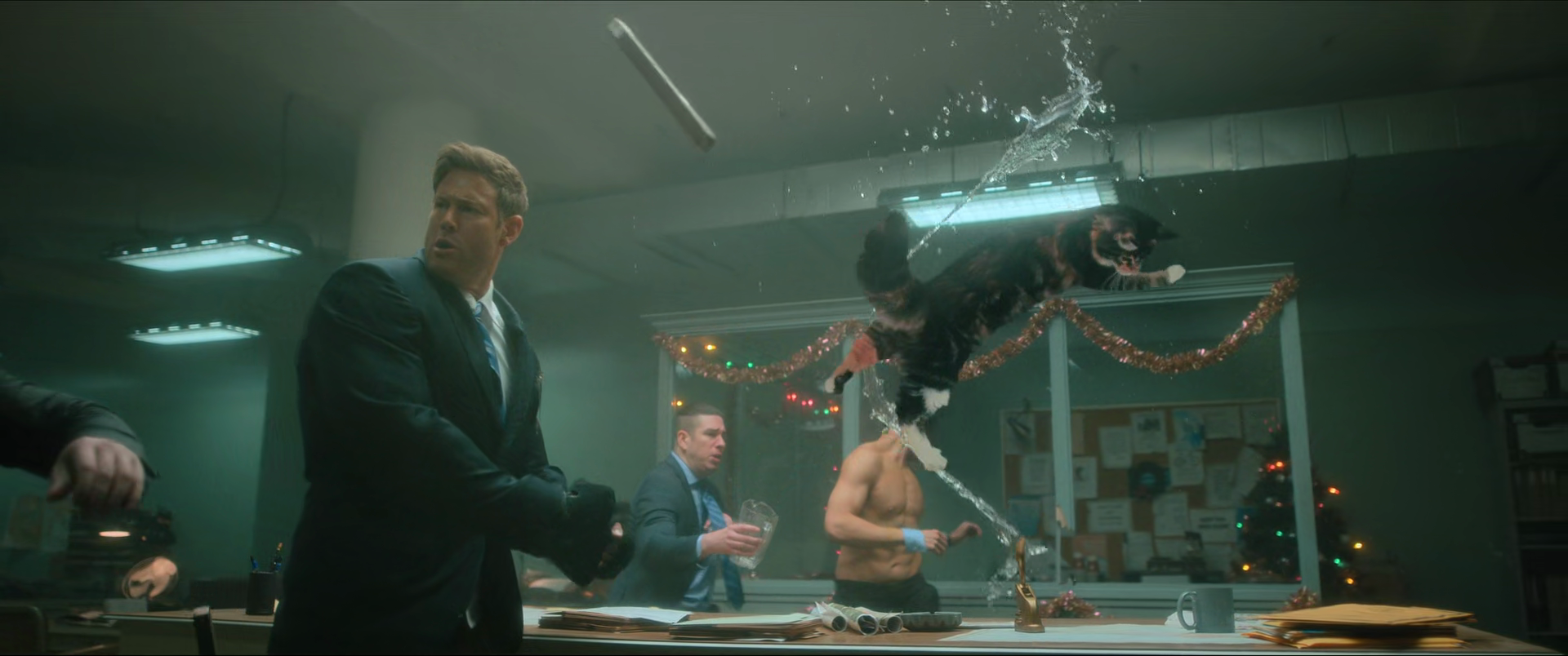 A chaotic scene in an office with a man in a suit looking shocked as a dog leaps off a table, spraying water from a hose.