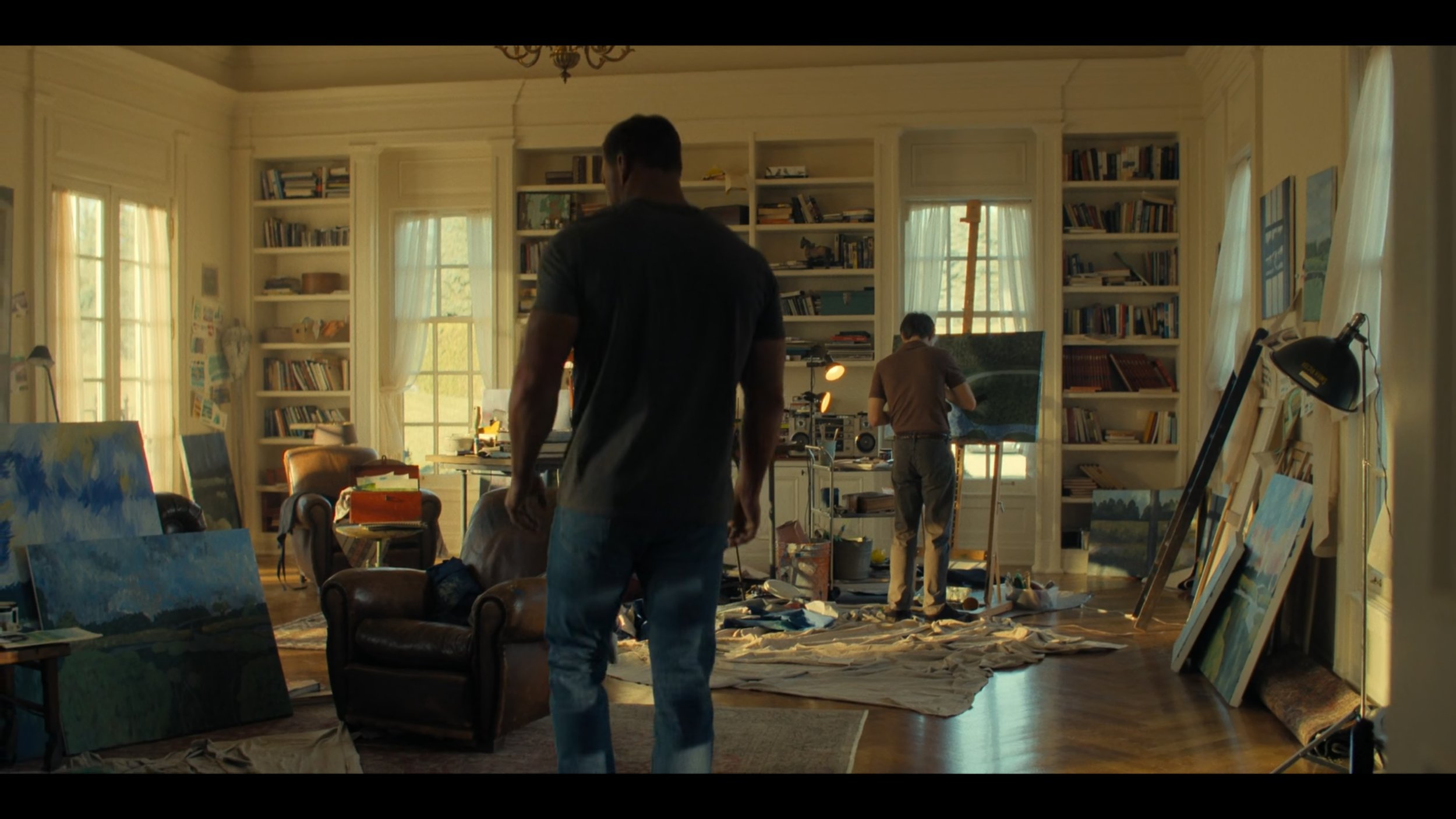 A spacious art studio with large windows, two men working on paintings, canvases on the floor and easels, and shelves filled with books and art supplies.