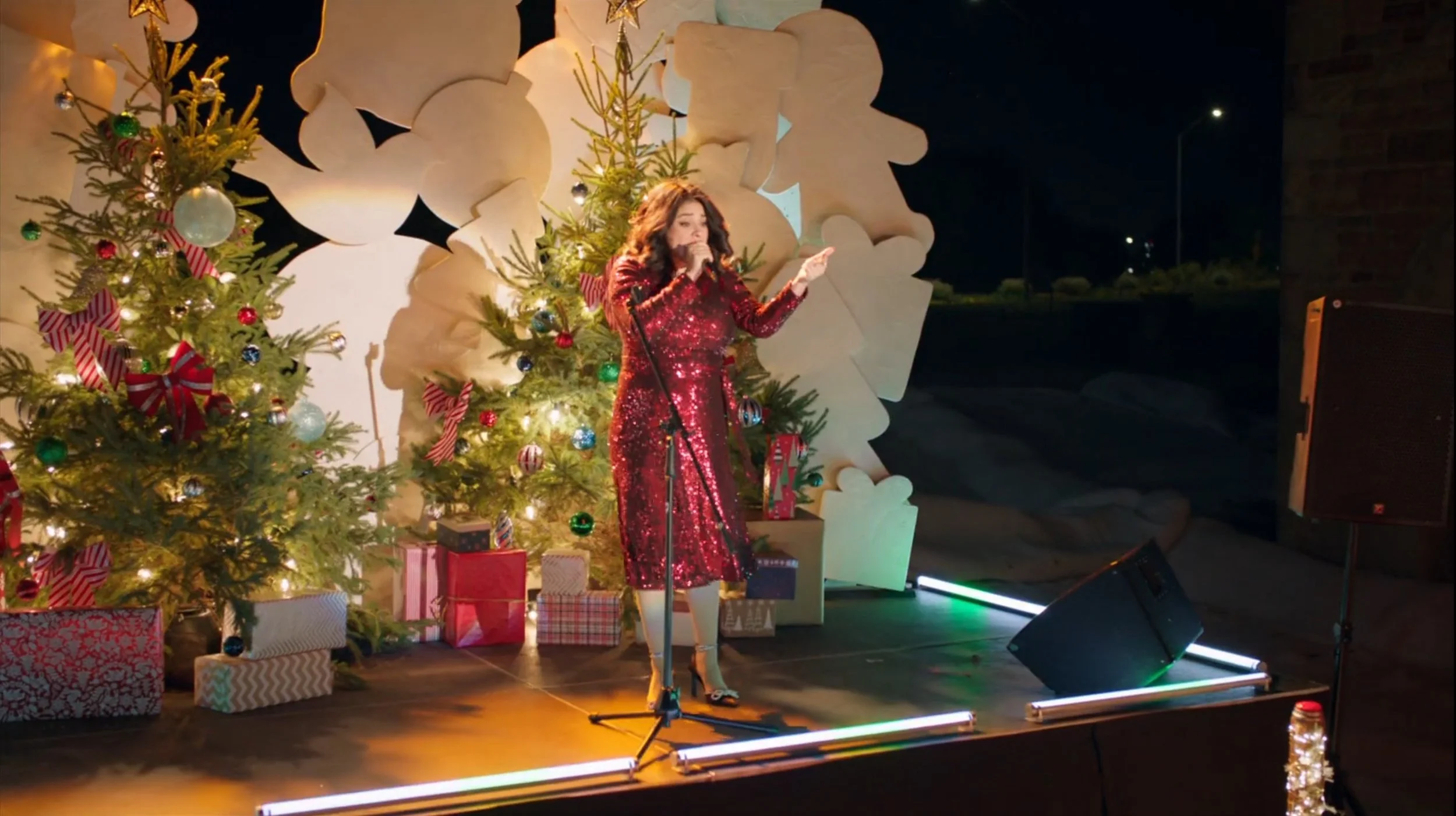 Woman singing on a decorated Christmas stage with Christmas trees and wrapped presents, at night.
