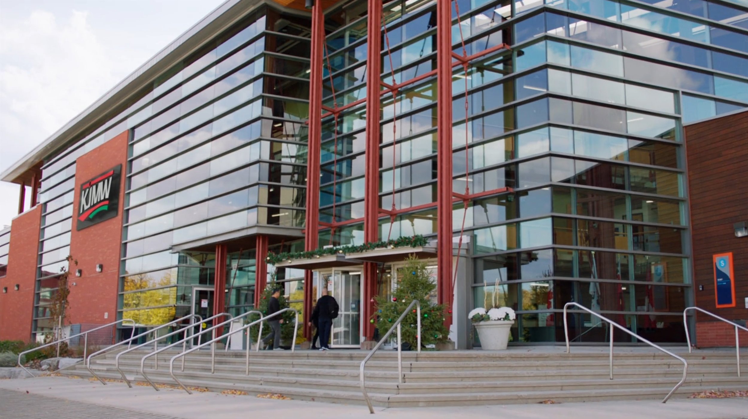 Exterior of a modern glass building with the KIWU logo on the side, outside stairs leading to the entrance, and people entering the building during daytime.