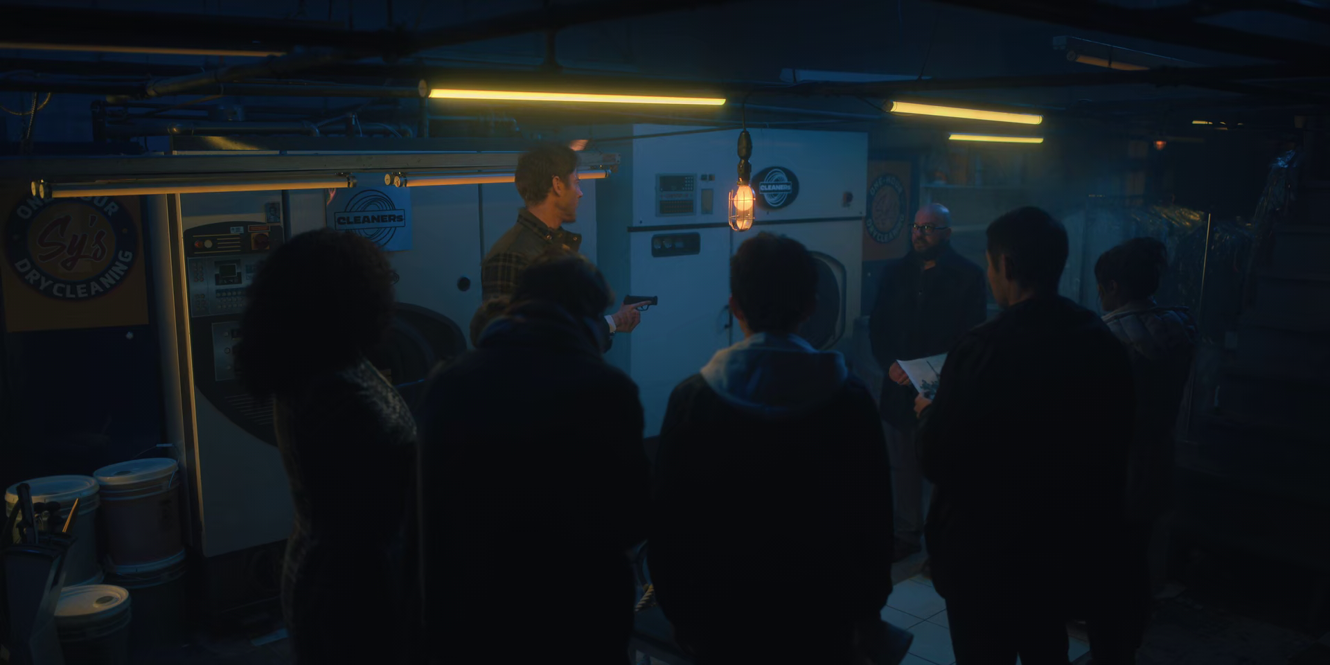 A small group of people in a dark laundry room, with two washers or dryers in the background, listening to a man speaking and holding a gun, with one person holding a paper.