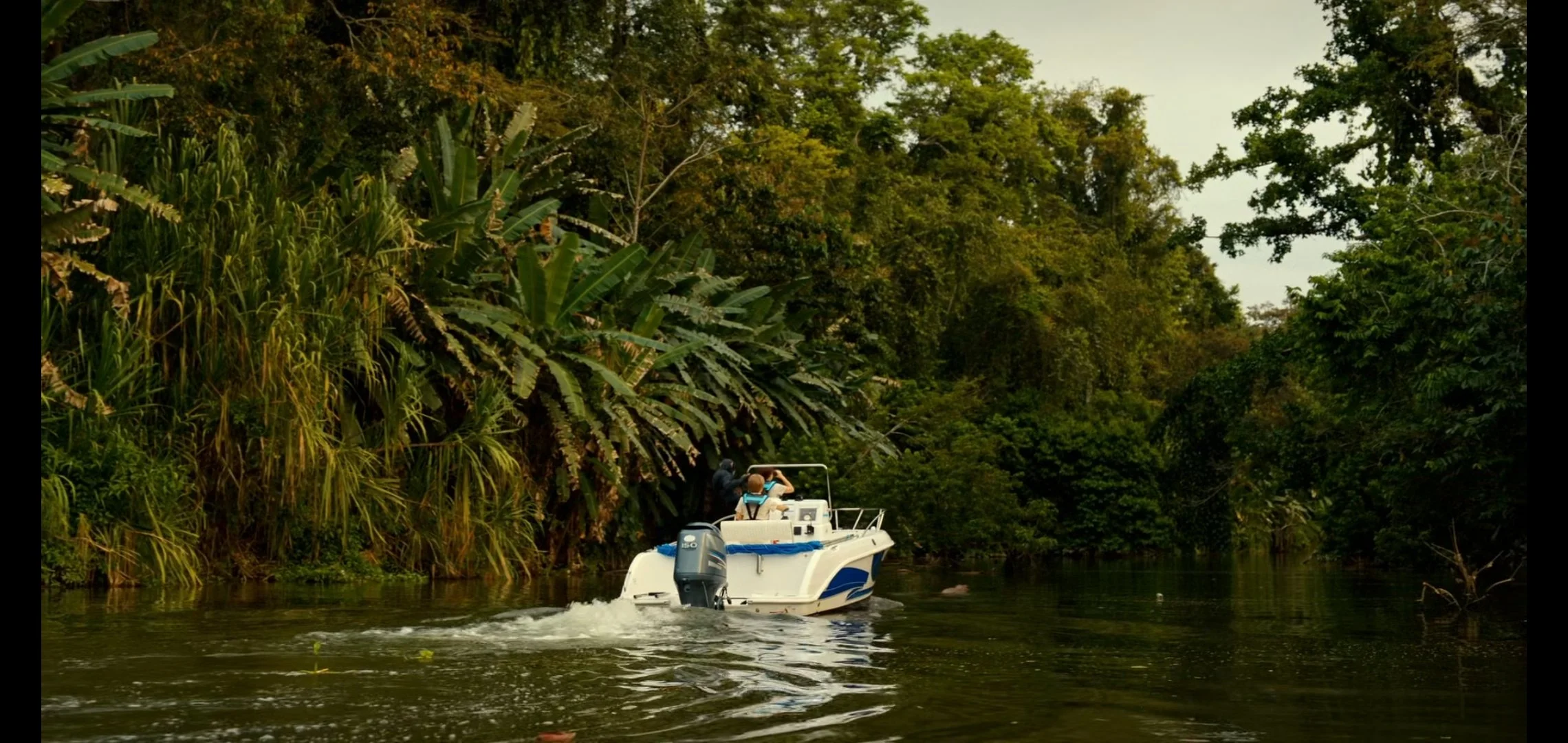 A small white motorboat with two people on board is cruising down a narrow, calm river surrounded by dense green foliage and trees.