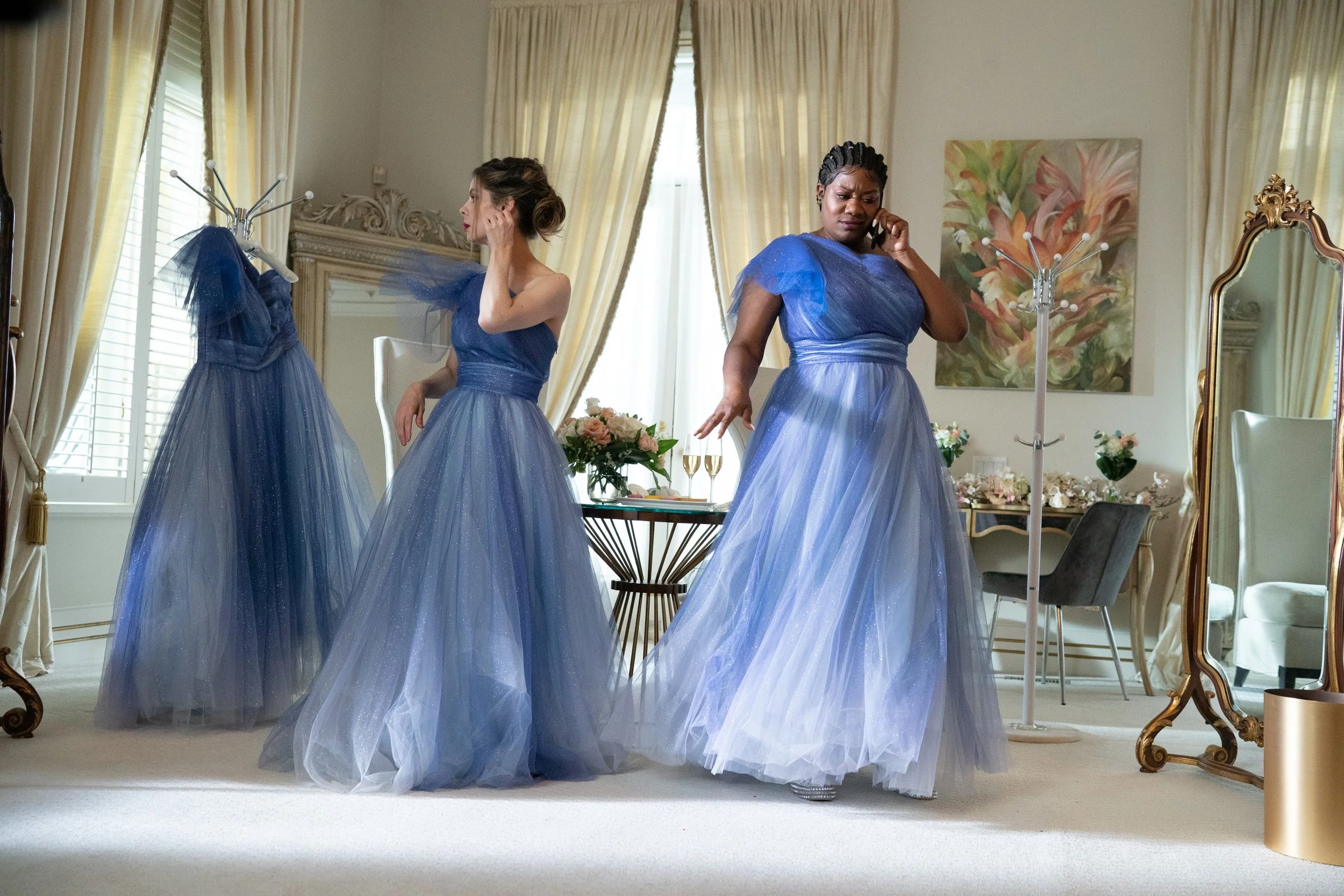 Three women in matching blue tulle gowns preparing for a formal event in a bright, elegantly decorated room with large windows, floral artwork, and gold-accented furniture.