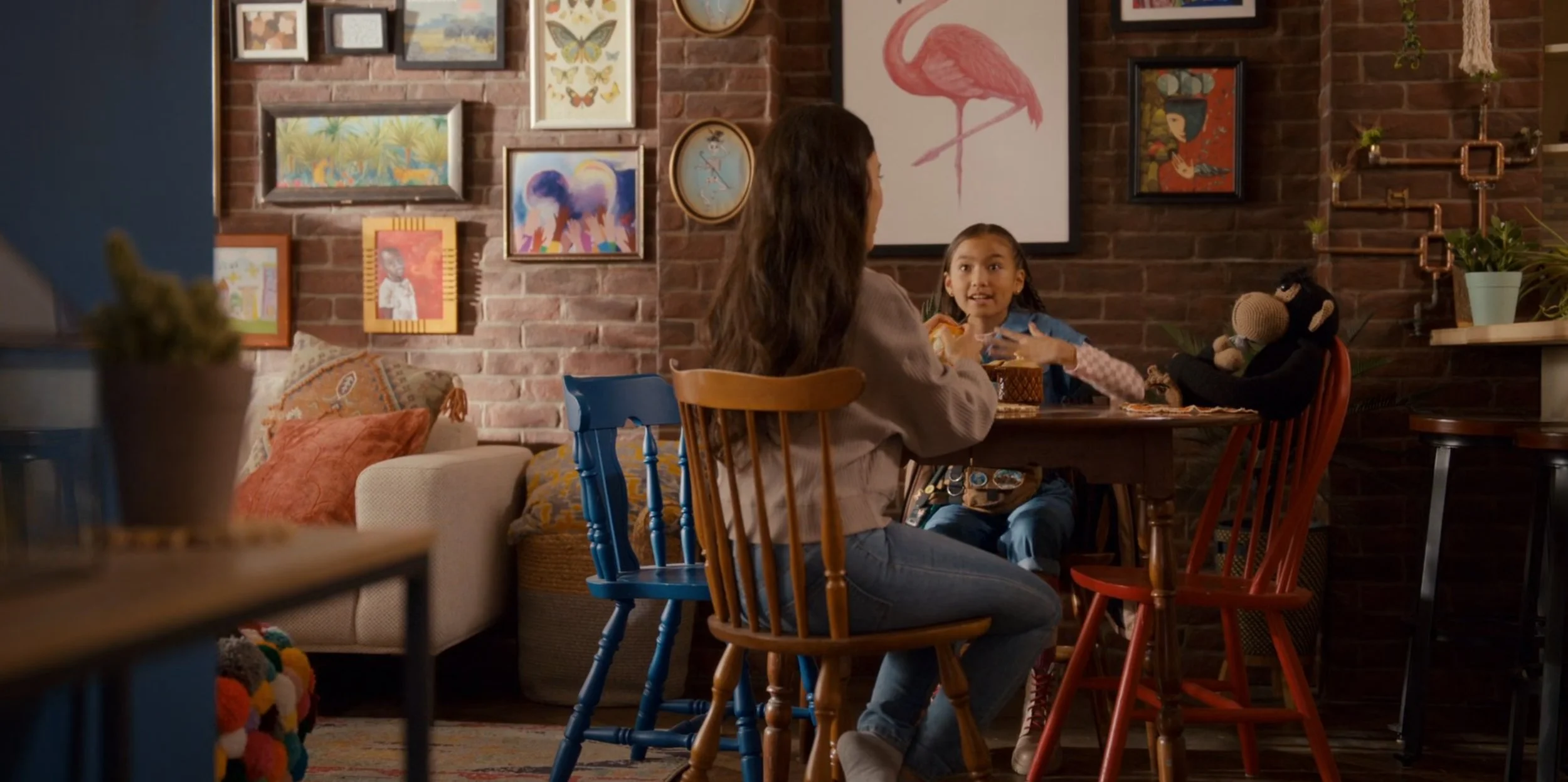 A woman and a girl are sitting at a round wooden table in a cozy cafe with a brick wall decorated with various framed artworks, including a flamingo, butterflies, and colorful paintings. The girl appears surprised or excited, while the woman is turned towards her, engaging in conversation. There are plush pillows on a white sofa in the background and a doll sitting at the table.
