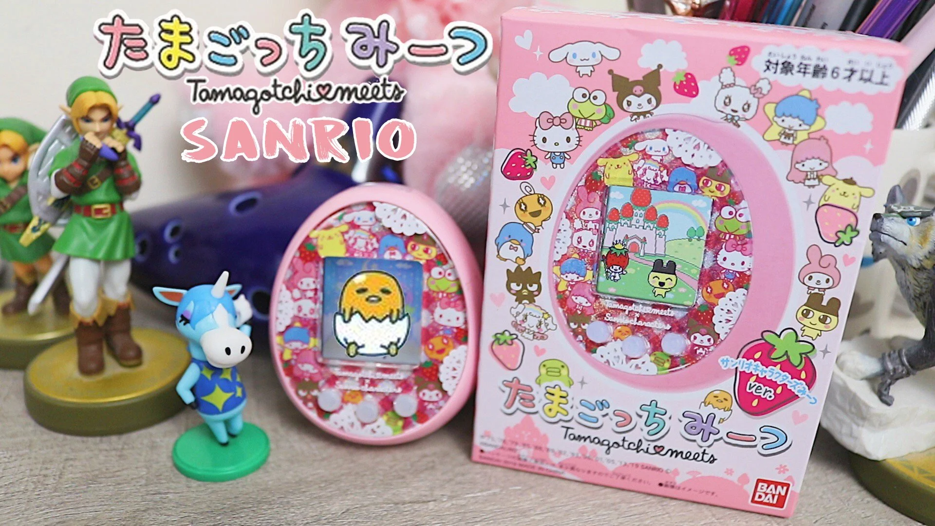 A Tamagotchi virtual pet device decorated with Sanrio characters. The device and packaging are pink with colorful Sanrio character illustrations. There are Nintendo amiibo figures of Link and a unicorn standing beside the Tamagotchi device.