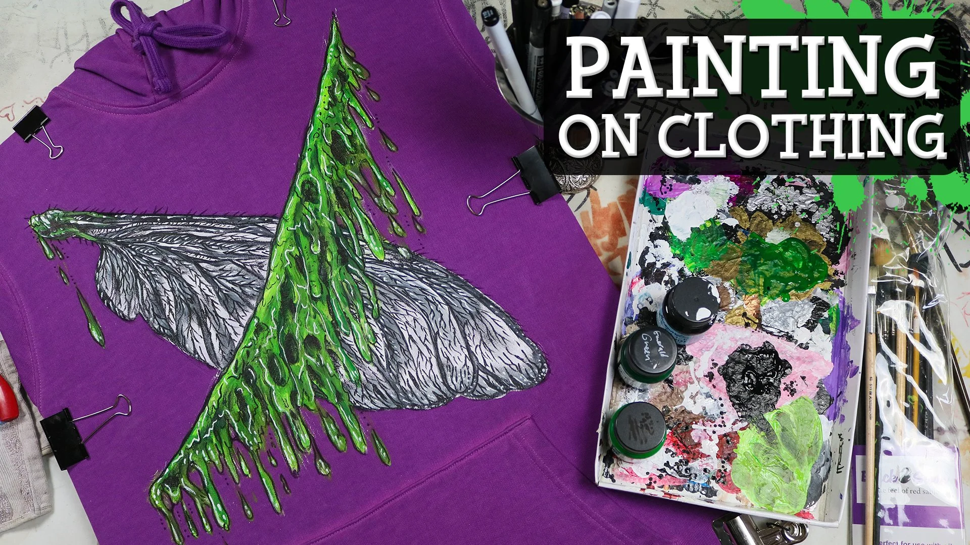 A purple hoodie with a hand-painted graphic of a black and white insect with green dripping paint on it, alongside painting supplies including paint jars, brushes, and a palette with various colors, and a black overlay with white text reading 'Painti