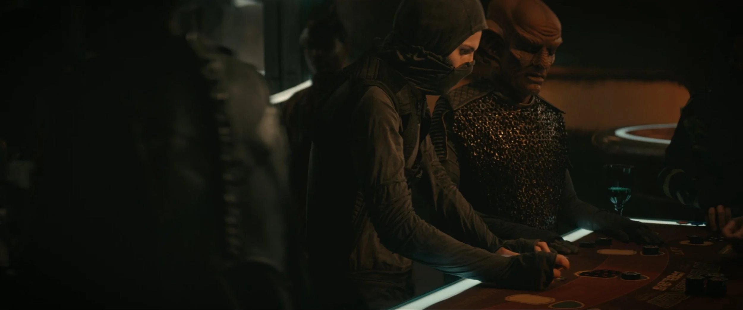 People in dark clothing playing poker at a casino table in a dimly lit room.