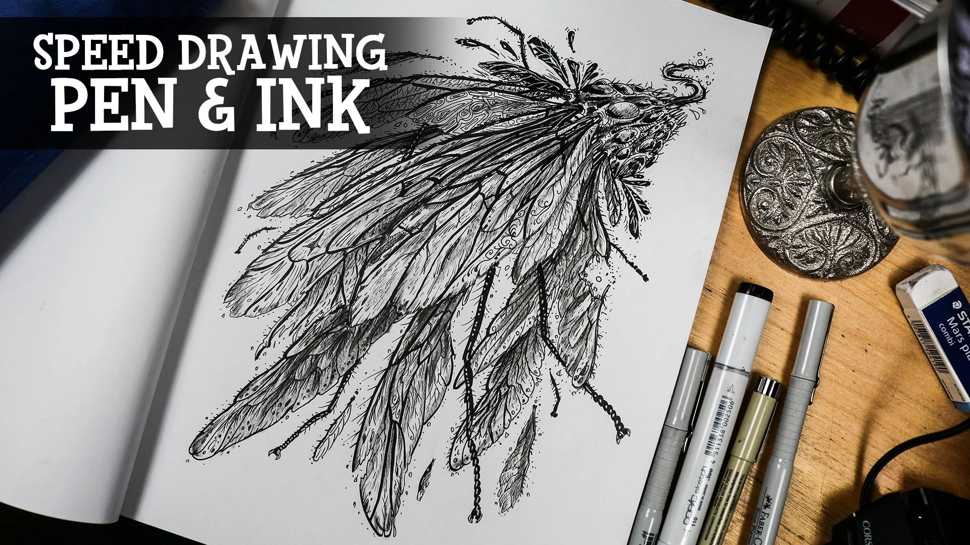 A detailed black and white ink illustration of a fantastical insect with multiple wings, surrounded by various pens and art supplies on a wooden surface.