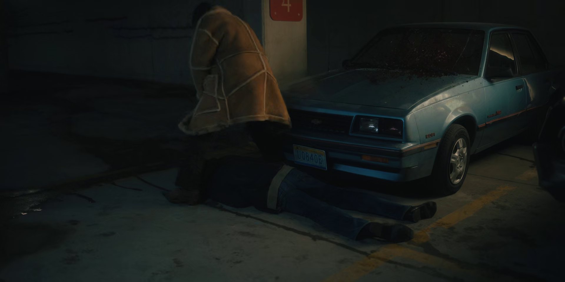 A person lying on the ground beneath a silver sports car in a dimly lit parking garage, with another person kneeling beside them. The person on the ground is wearing dark clothing and appears to be injured or unconscious.