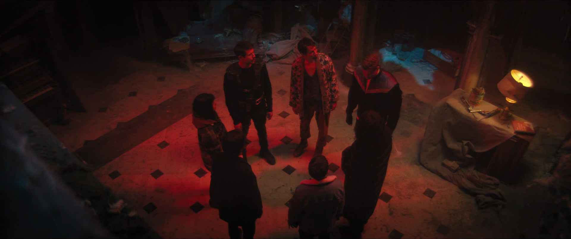 Group of people standing in a circle in a dimly lit room with red and blue lighting, some sitting on a sofa and others standing around.
