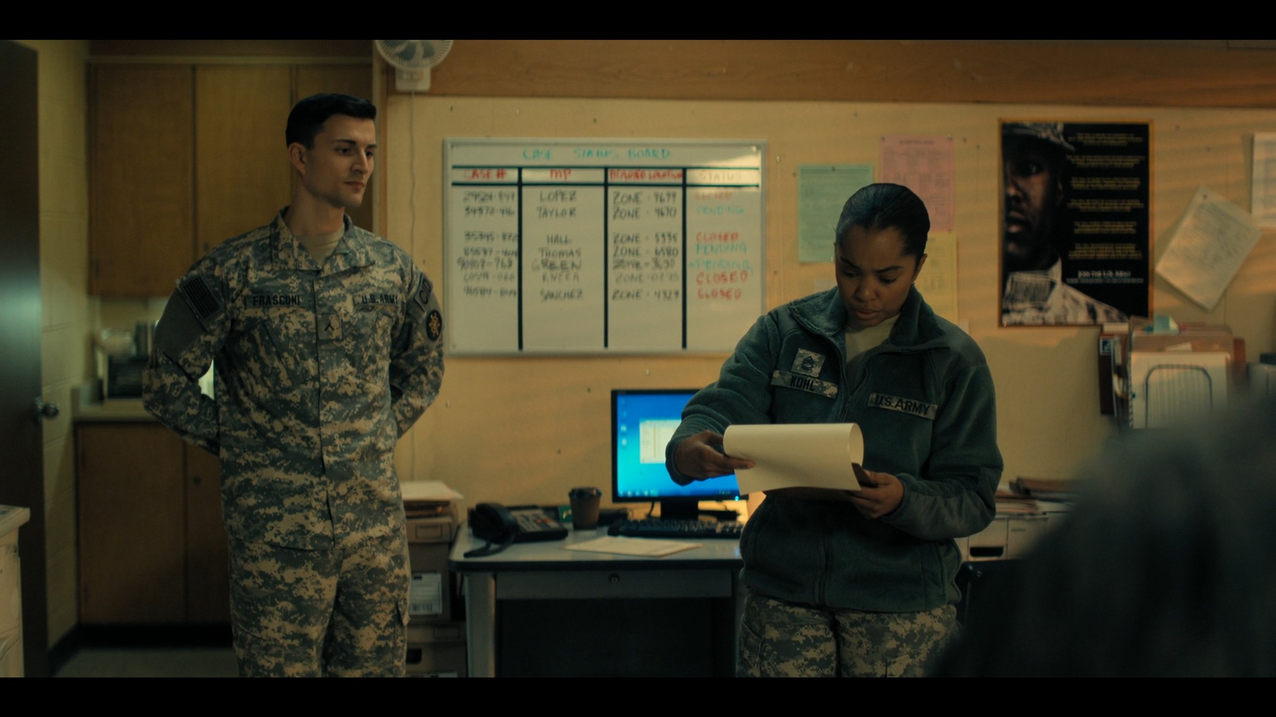 A male soldier in camouflage uniform stands with hands behind his back, looking at a female soldier in a U.S. Army uniform, who is reading a notebook in an office setting with a computer, desk, and bulletin board in the background.