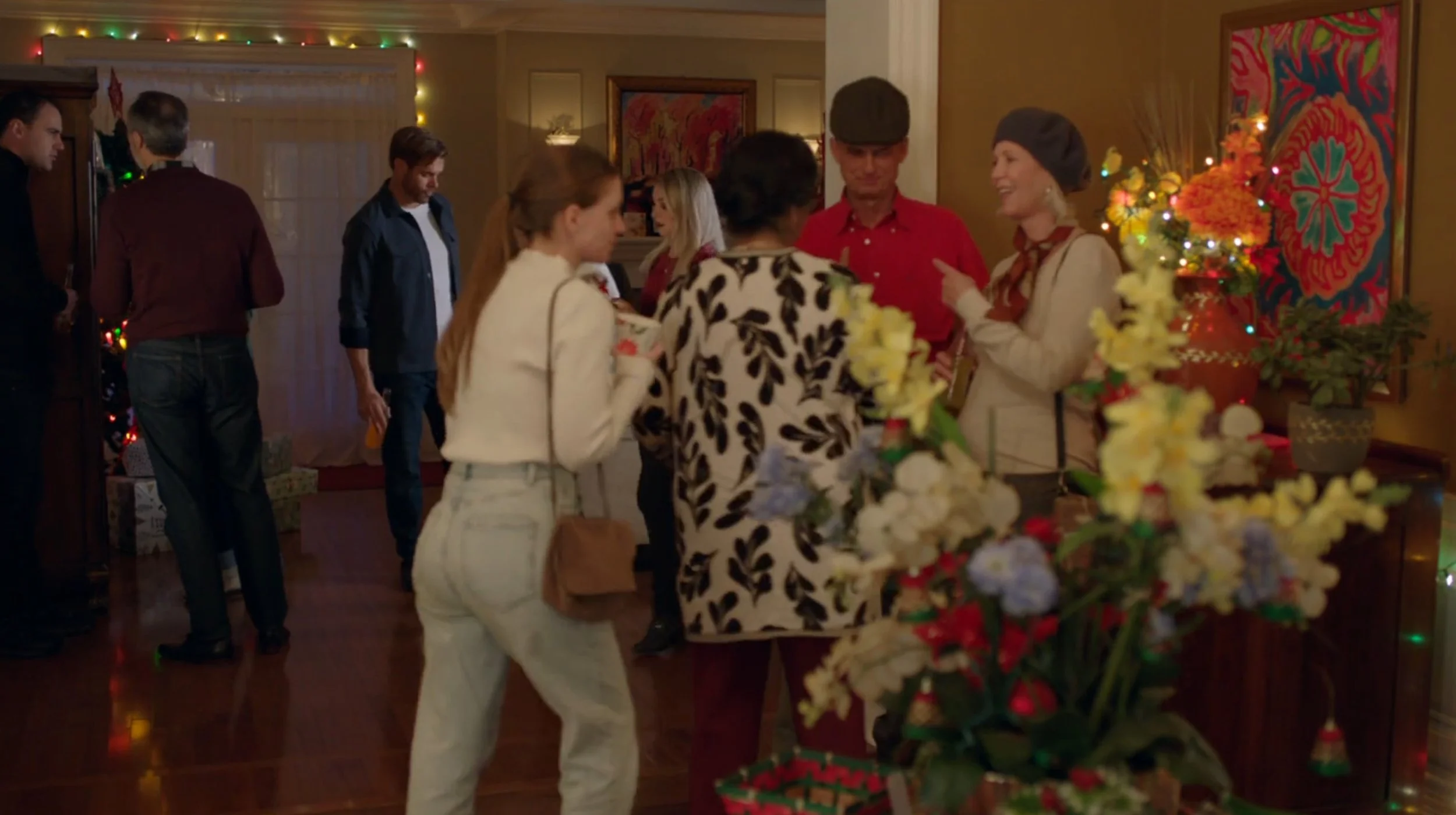 People socializing at a Christmas gathering in a warmly decorated living room with colorful artwork, string lights, and floral arrangements.
