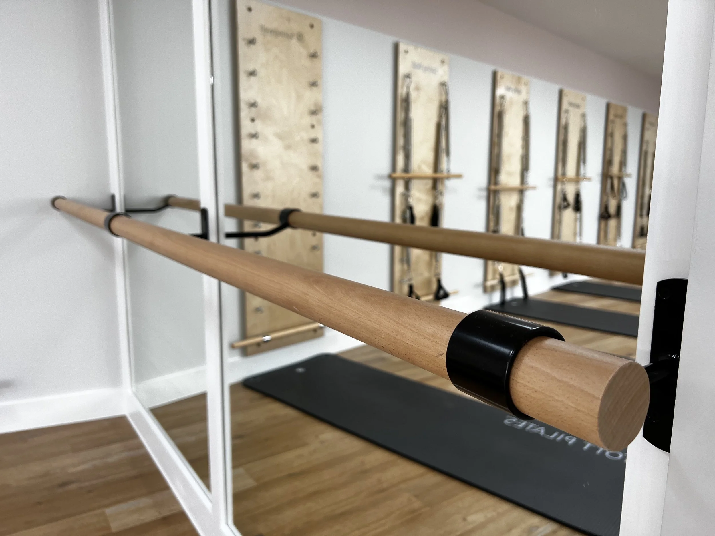 Yoga studio interior with wall-mounted pilates equipment, a wooden barre, and reflection in a mirror, on a wooden floor.