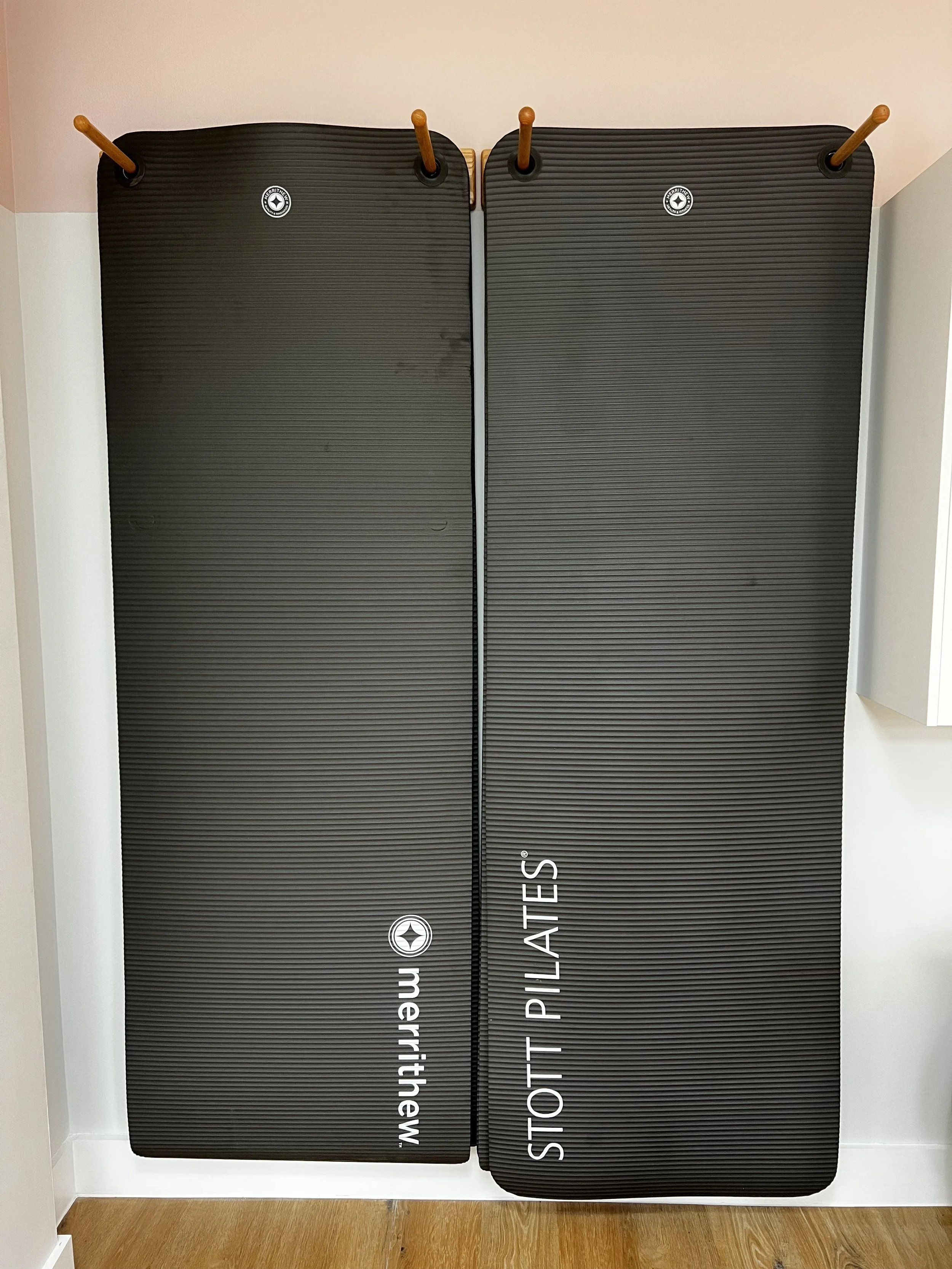 Two black pilates mats hanging vertically on wooden pegs against a light-colored wall, branded with "Merrithew" and "STOTT PILATES."