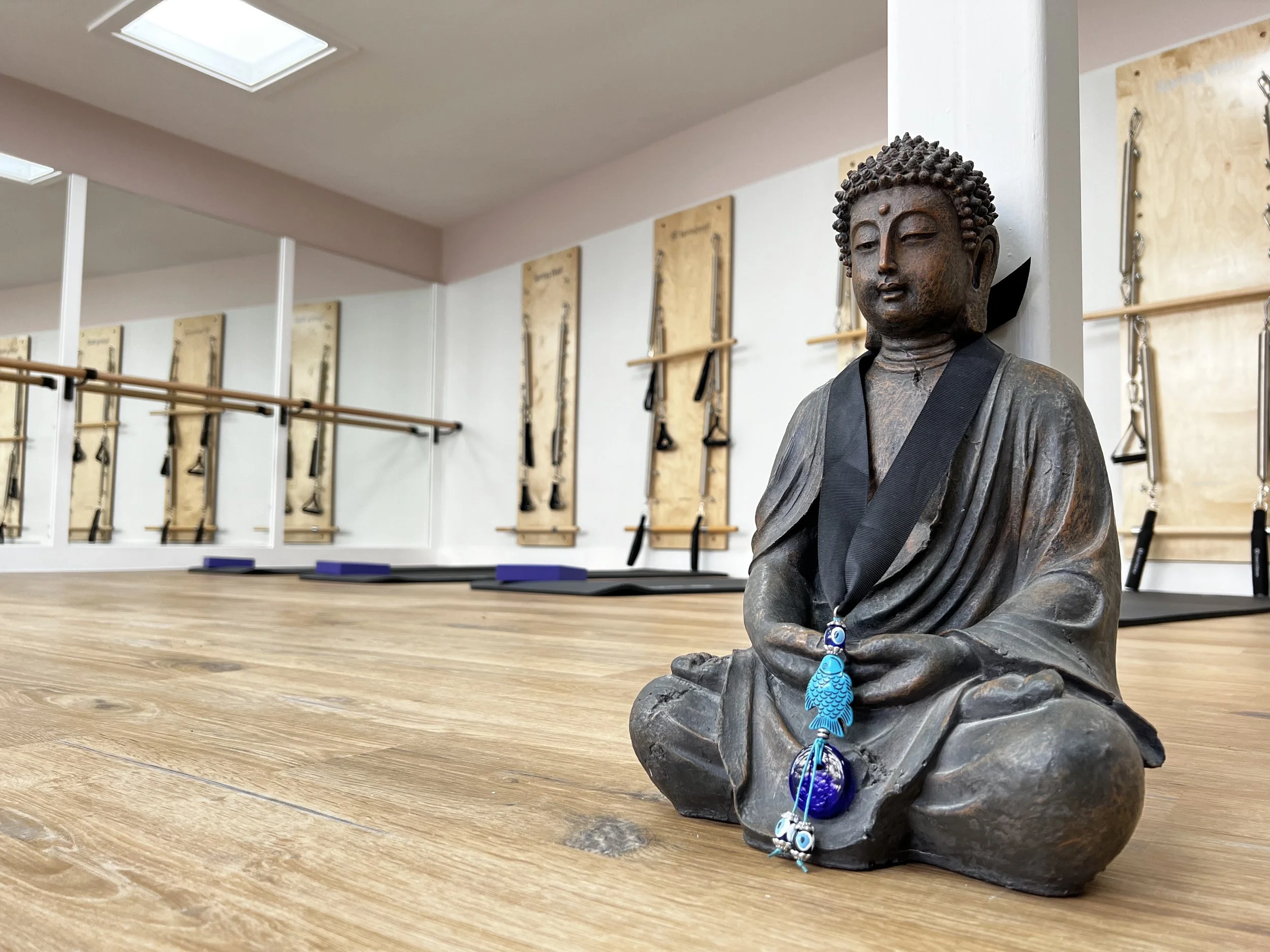Yoga studio with a Buddha statue, wooden floor, wall mirrors, Pilates equipment, and exercise mats.