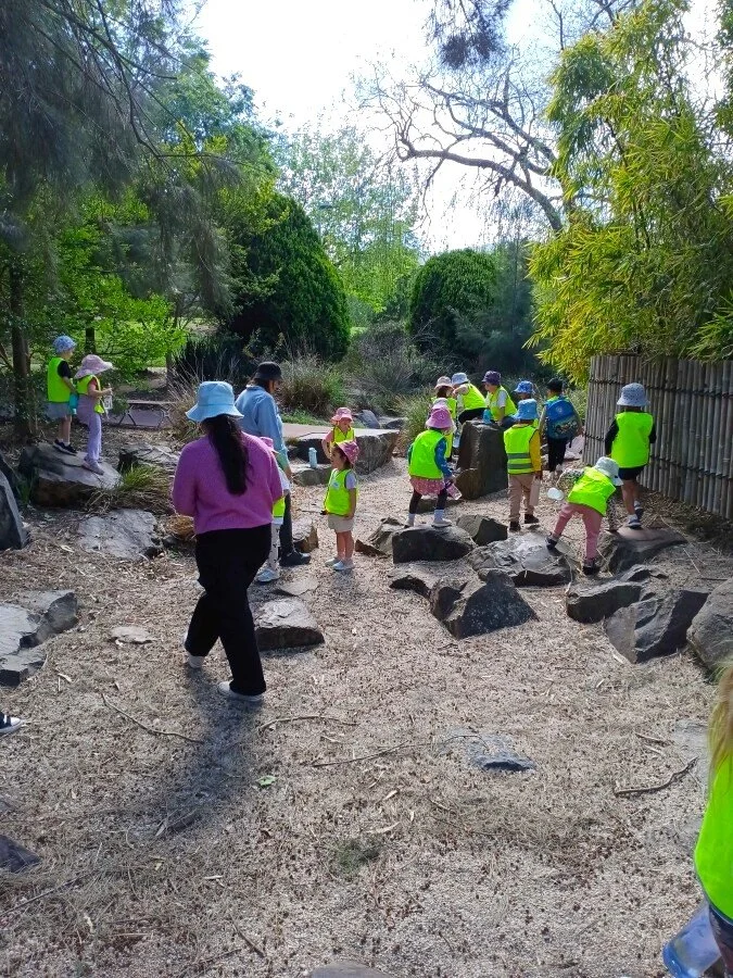 A group of children and adults on a nature walk in a forested area, some wearing bright yellow safety vests and hats, exploring rocks and the natural landscape.