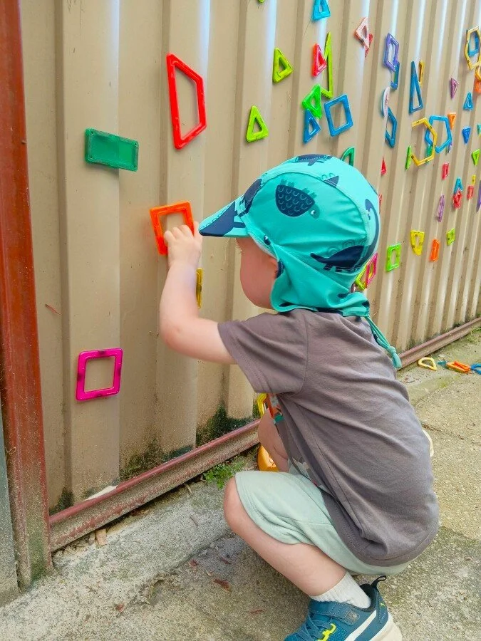 A young boy crouches and places a colorful geometric shape onto a metal fence decorated with more similar shapes in bright colors.