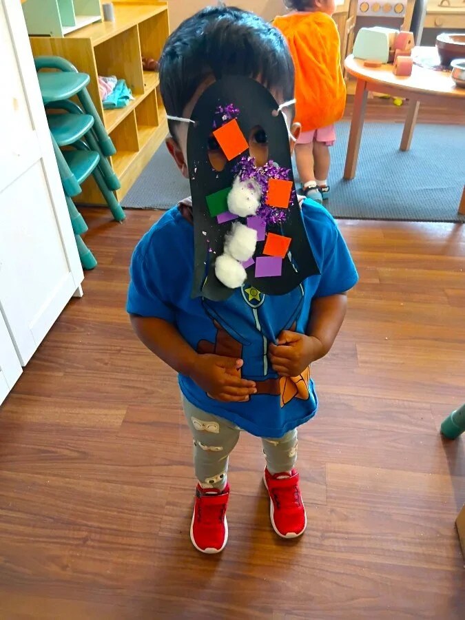 A young child standing indoors on wooden flooring, wearing a blue shirt and red sneakers, with a homemade craft mask covering their face. The mask is decorated with colorful foam shapes, cotton balls, purple glitter, and held up with a string around 