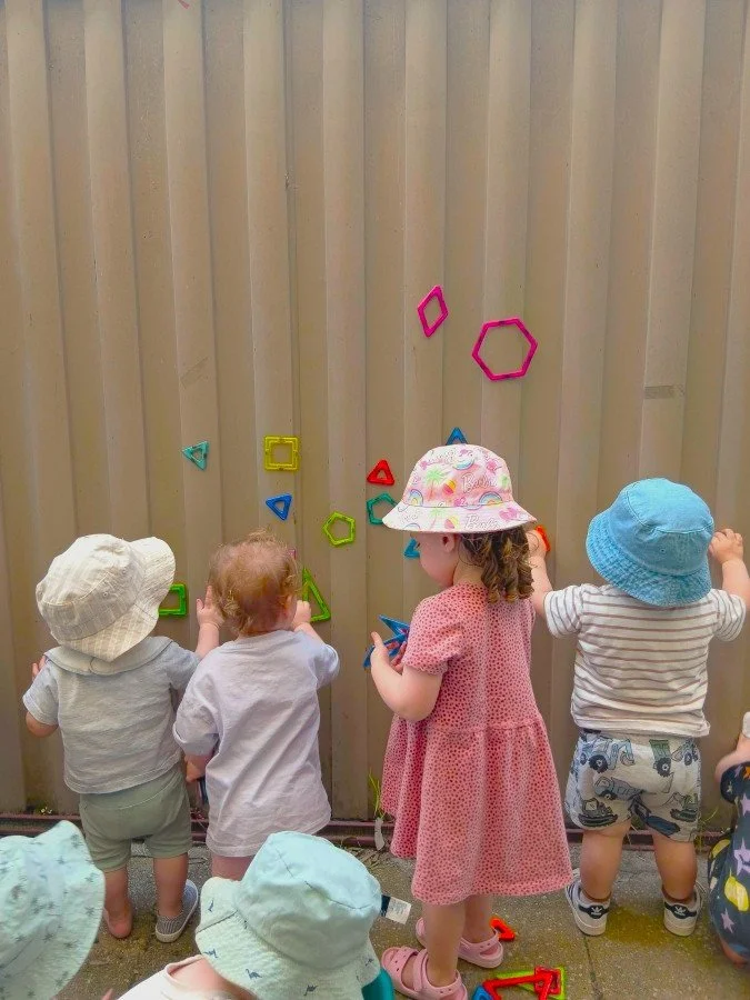 Four young children with hats building with colorful magnetic shapes on a wooden fence.