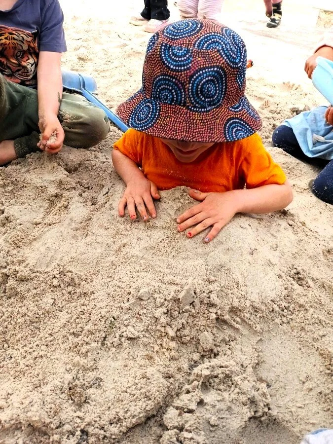 A child wearing an orange shirt and a colorful patterned sun hat is lying on their stomach in the sand, with their hands in the dirt, at the beach. Other children are sitting or playing nearby.
