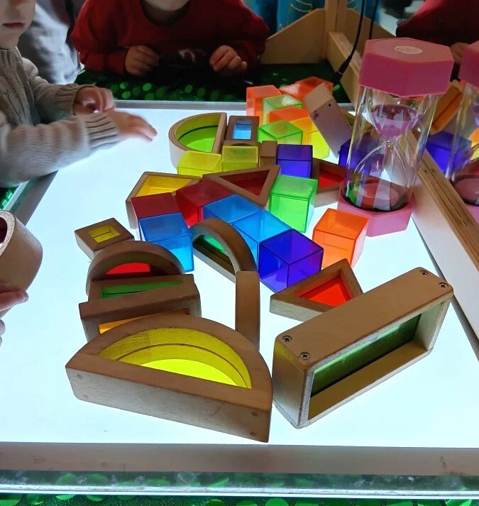 Children's educational toys on a light table, including colorful geometric shapes, blocks, and magnifying glasses, in a classroom setting.