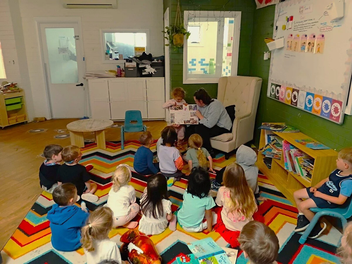 Teacher reading a book to a group of young children sitting on a colorful rug in a classroom. Children are attentively looking at the book, and the classroom has educational posters and books.