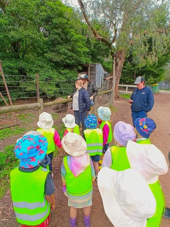 Group of young children wearing bright yellow safety vests and hats on a farm tour, looking at a person in a hat and long coat near a chicken coop.