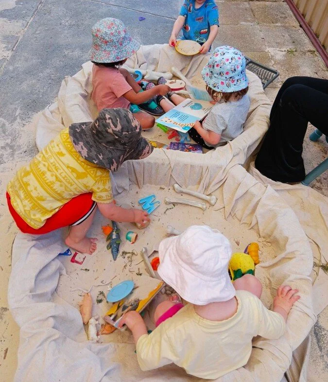 Children playing in a makeshift sandbox on a large fabric sheet, with some reading books and others playing with toys and sand.